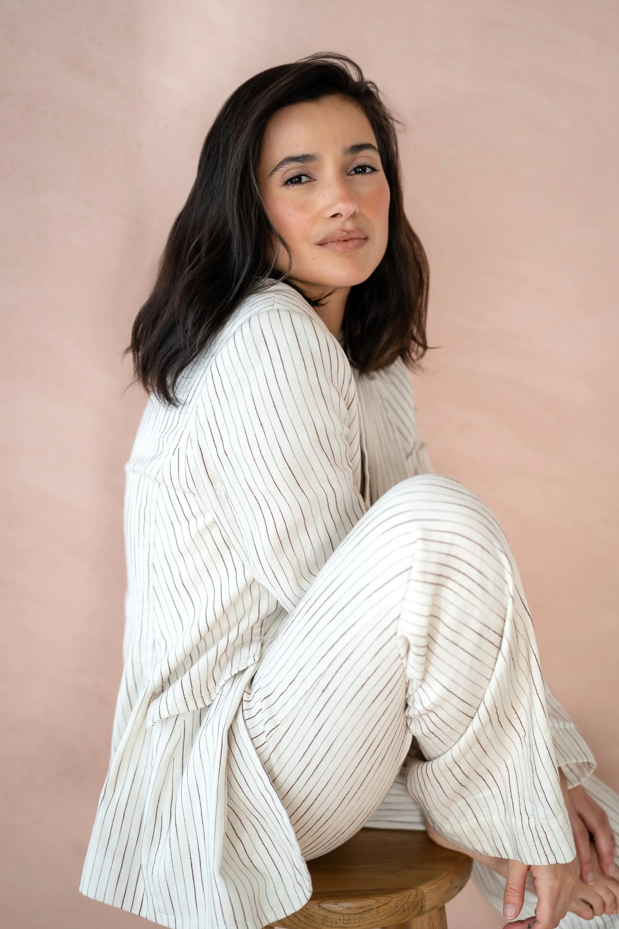A woman with dark hair and light skin, wearing a white striped outfit, sitting on a wooden stool against a light pink background.