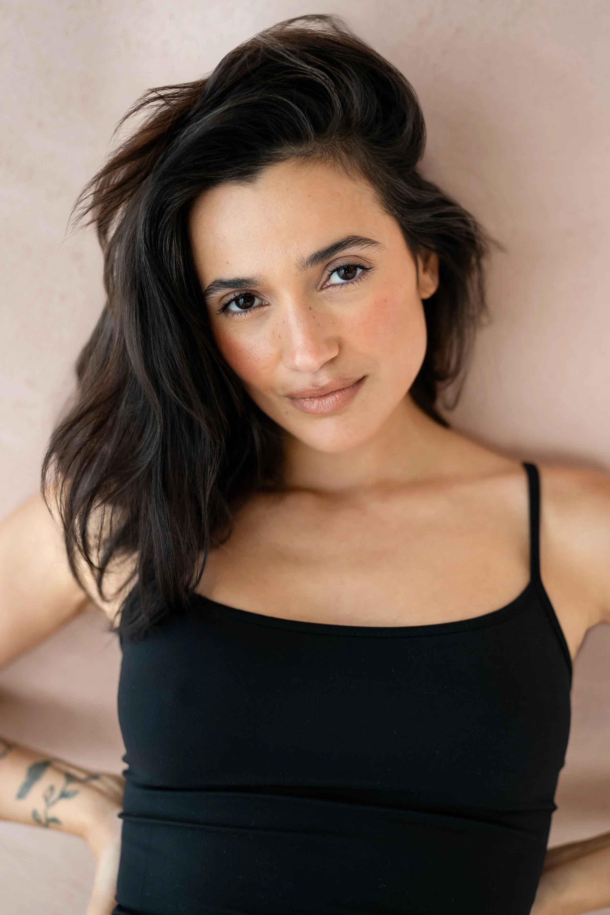 Close-up portrait of a woman with dark hair and a black tank top, looking at the camera with a soft smile, against a light-colored wall.
