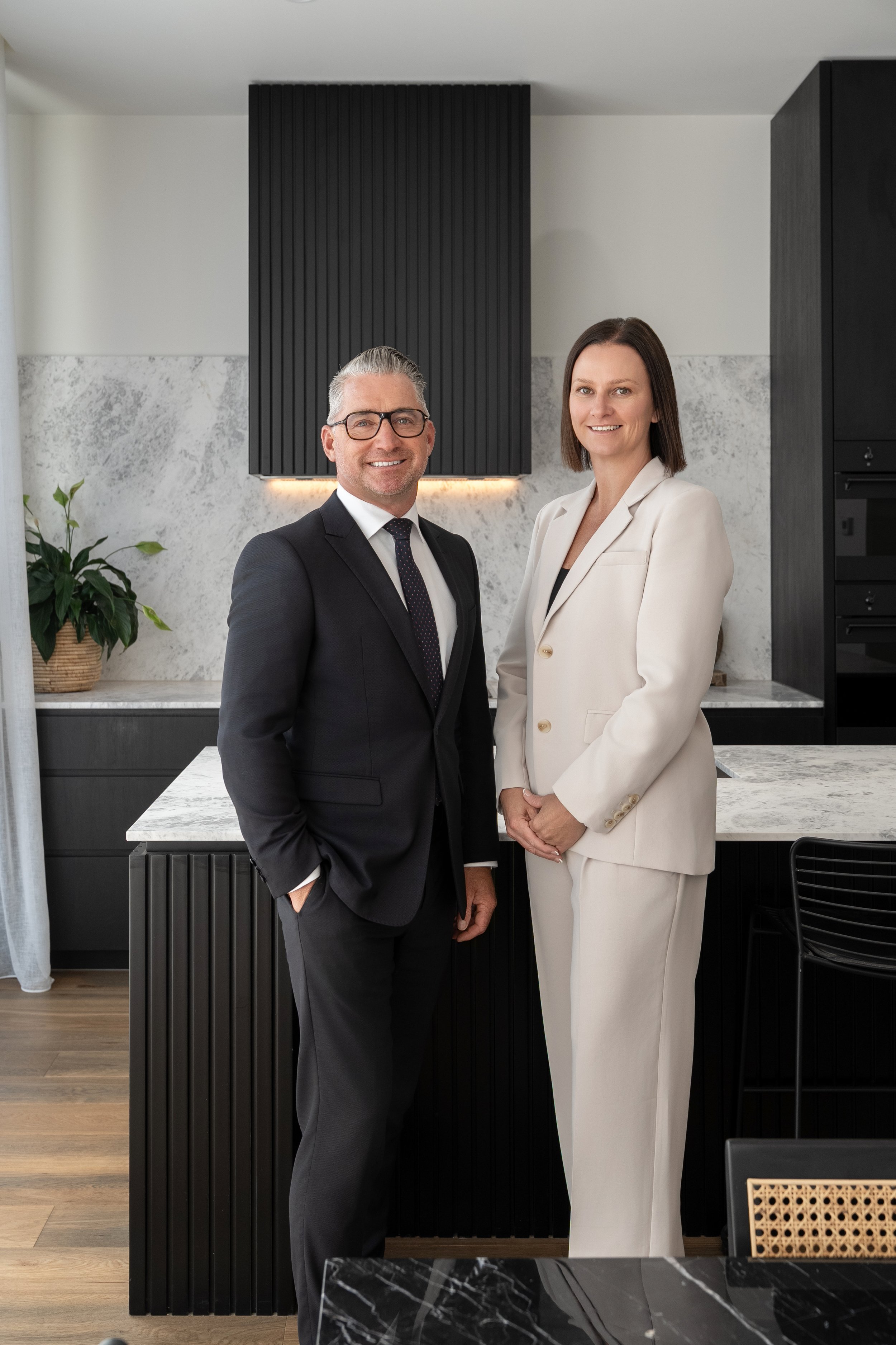 A man and woman dressed in business attire standing in a modern kitchen with black and white decor, smiling at the camera.