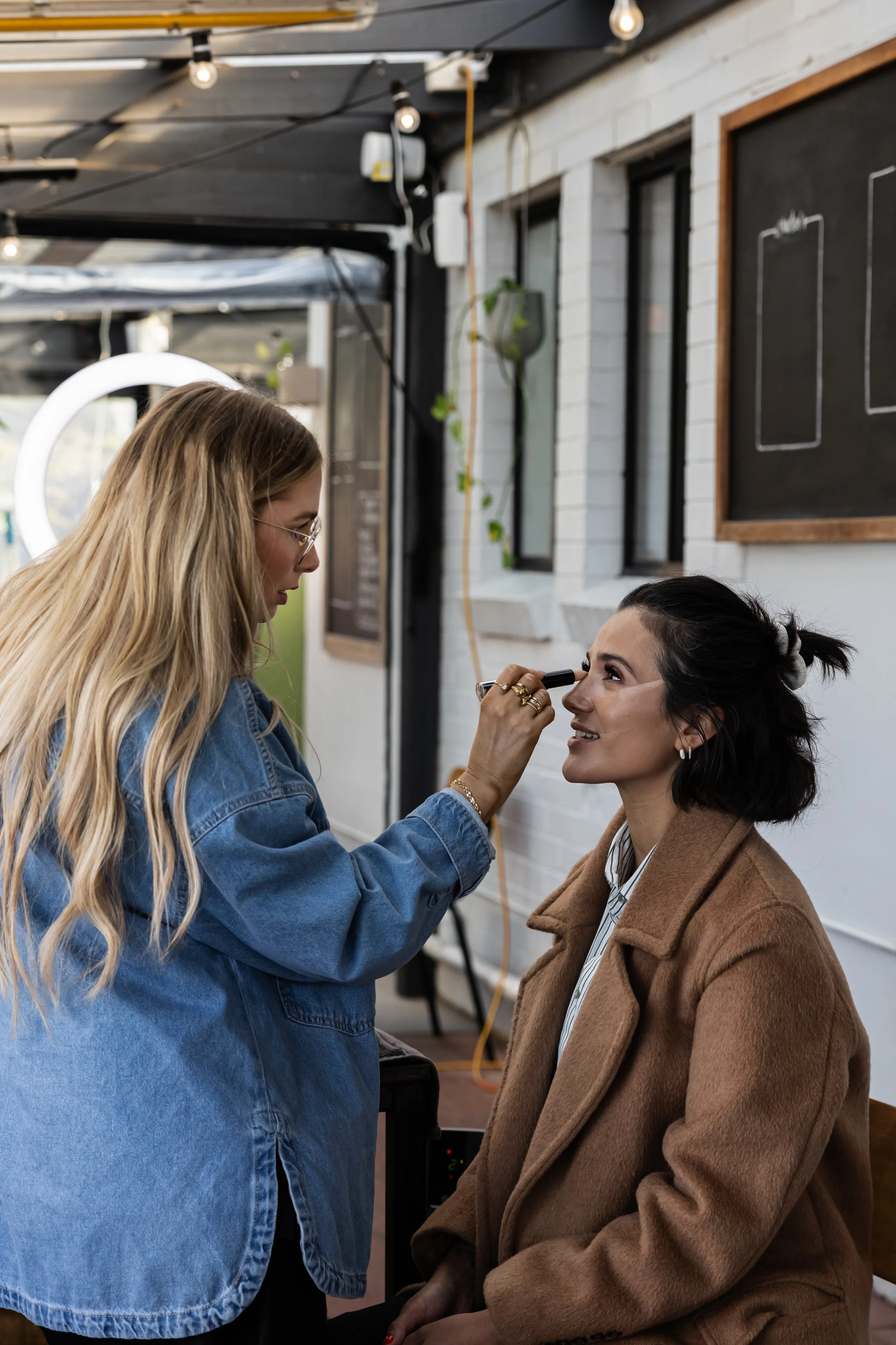 A makeup artist applies makeup to a seated woman with dark hair, wearing a brown coat, in a modern studio with blackboards, windows, and hanging lights.