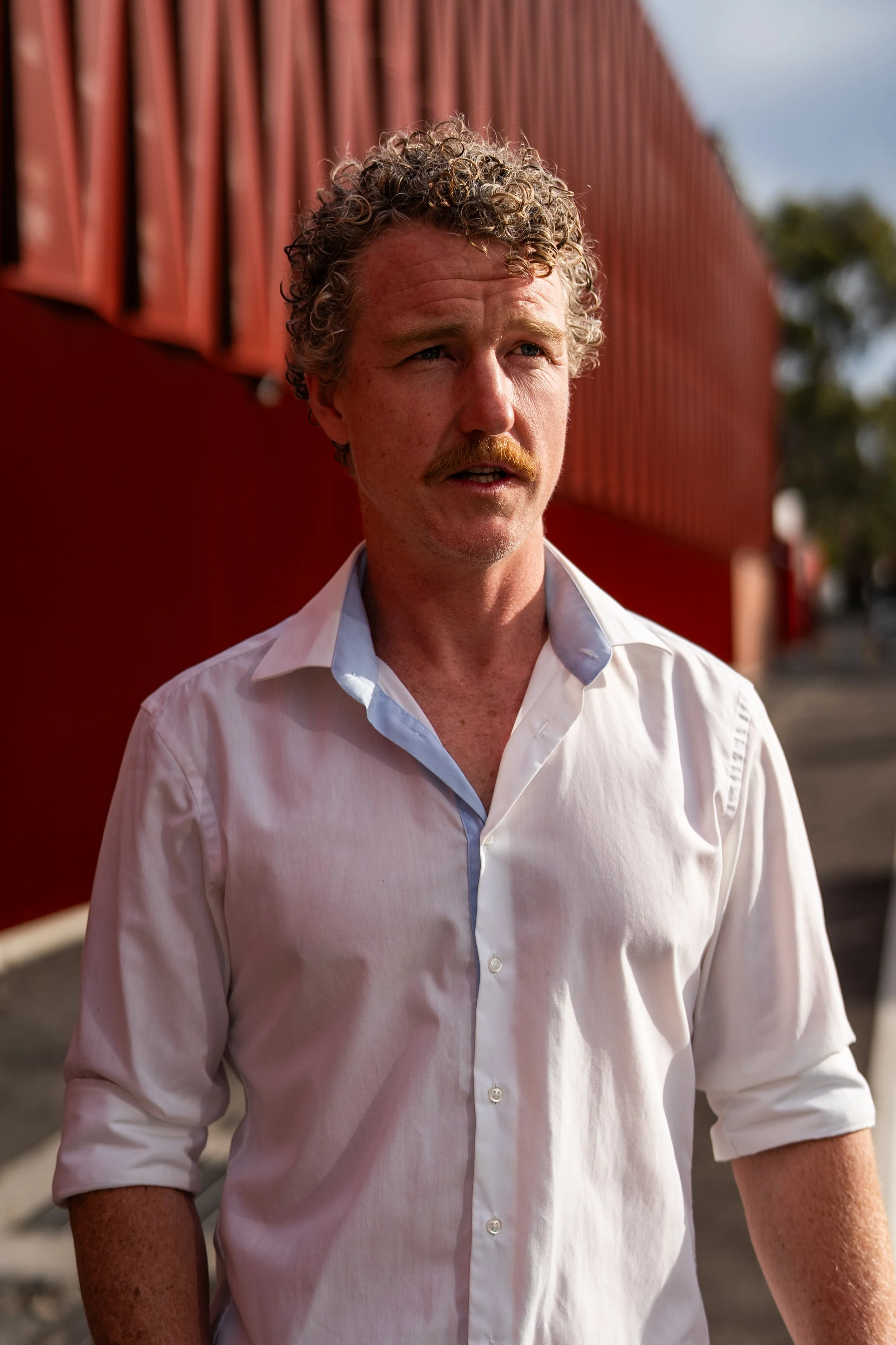A man with curly hair and a mustache wearing a white dress shirt in front of a red building.