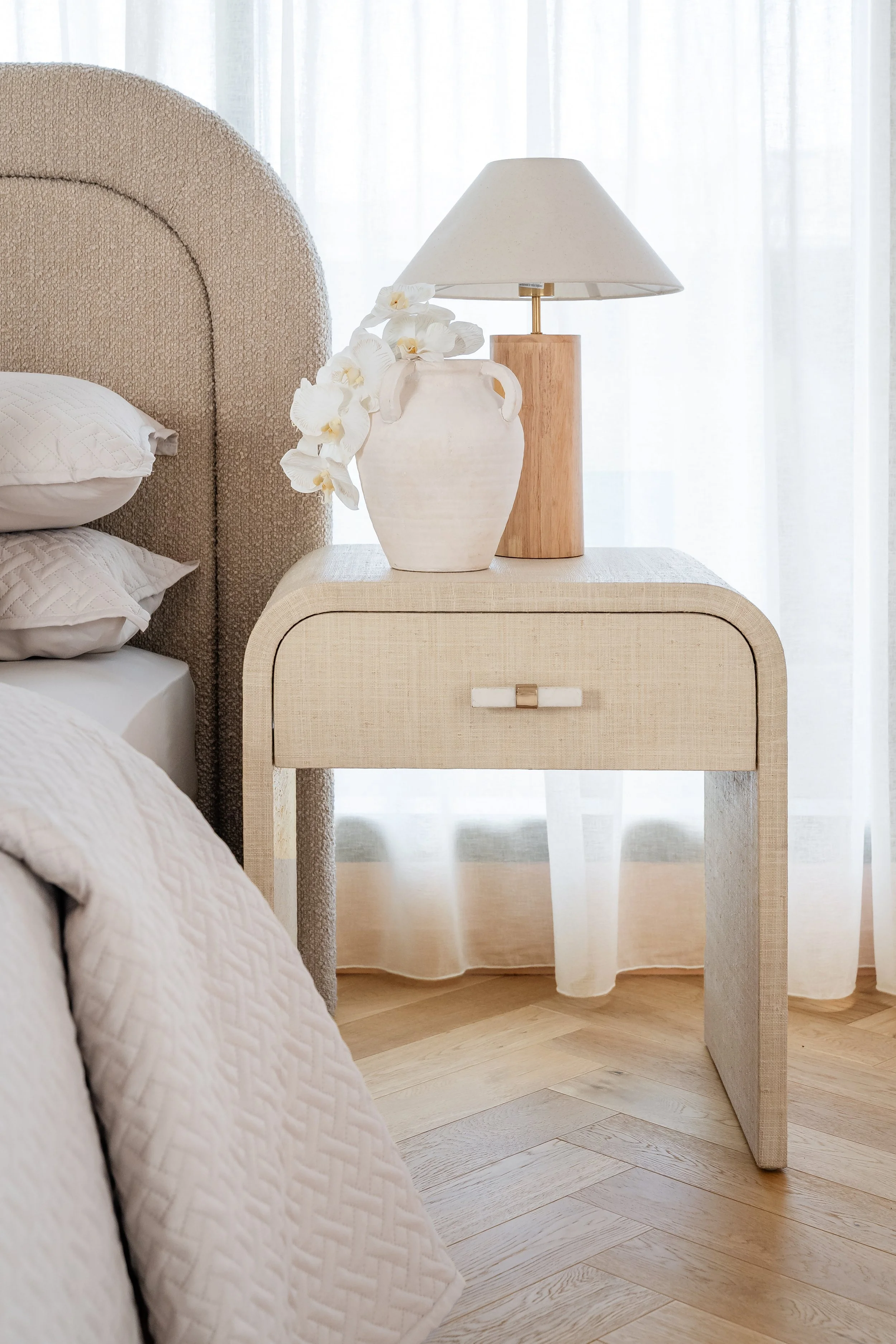 A bedroom nightstand with a lamp, a white vase of white orchid flowers, and a bed with white bedding against sheer white curtains and light wood flooring.