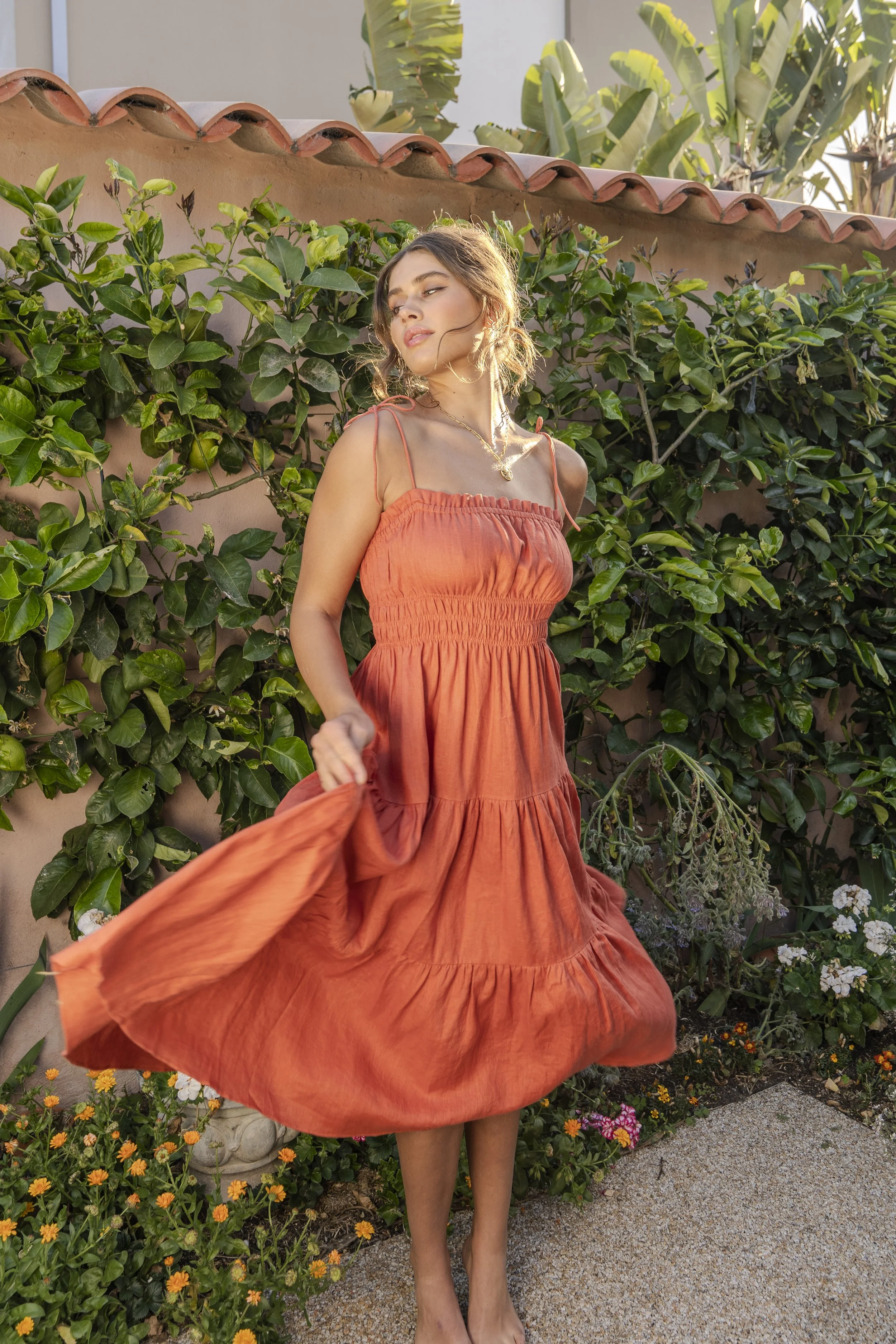 A woman in an orange dress standing in a garden with green plants and flowers.
