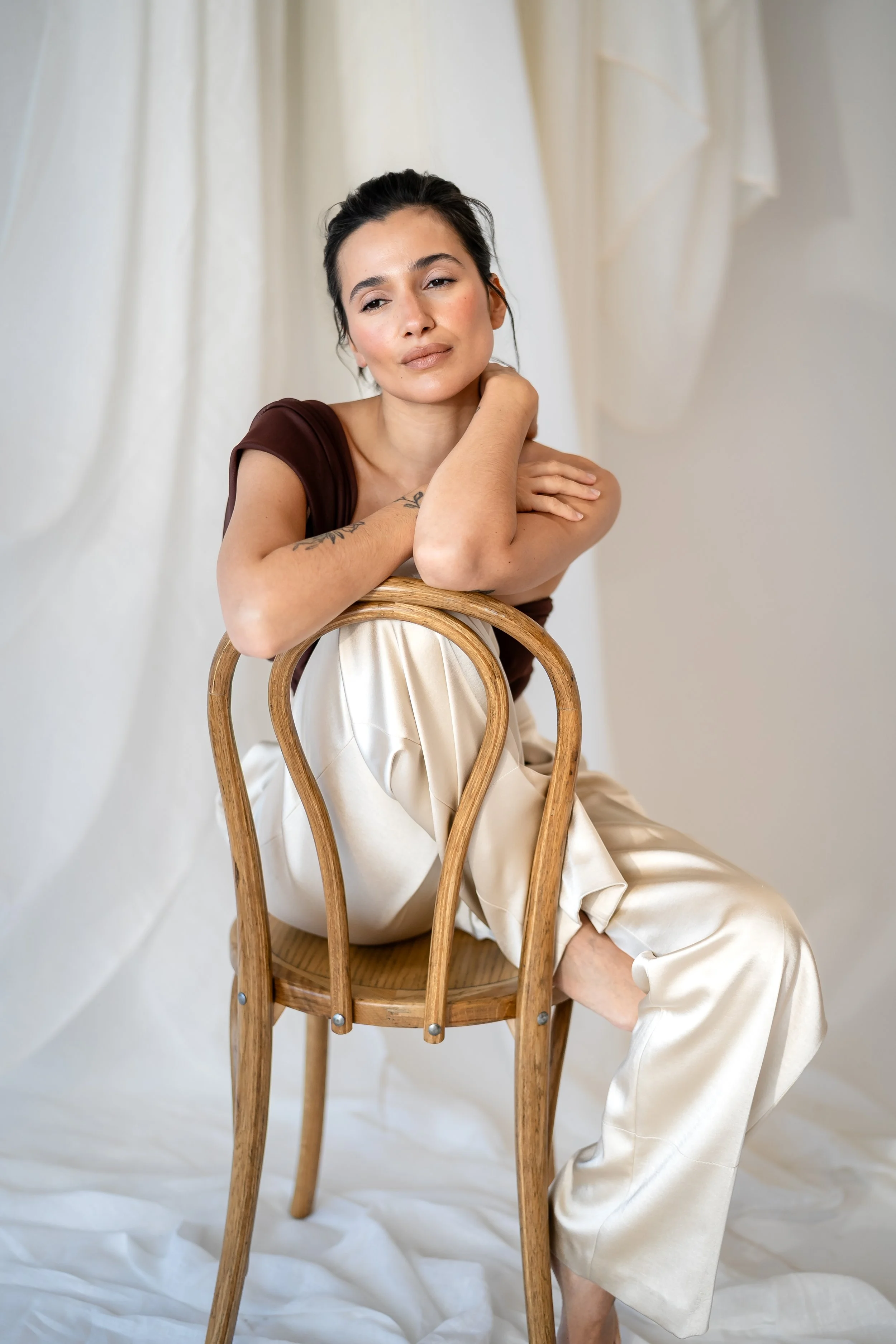 A woman with dark hair, wearing a dark brown top and cream satin pants, sitting on a wooden chair with her arms resting on the backrest and looking at the camera, against a light-colored backdrop.