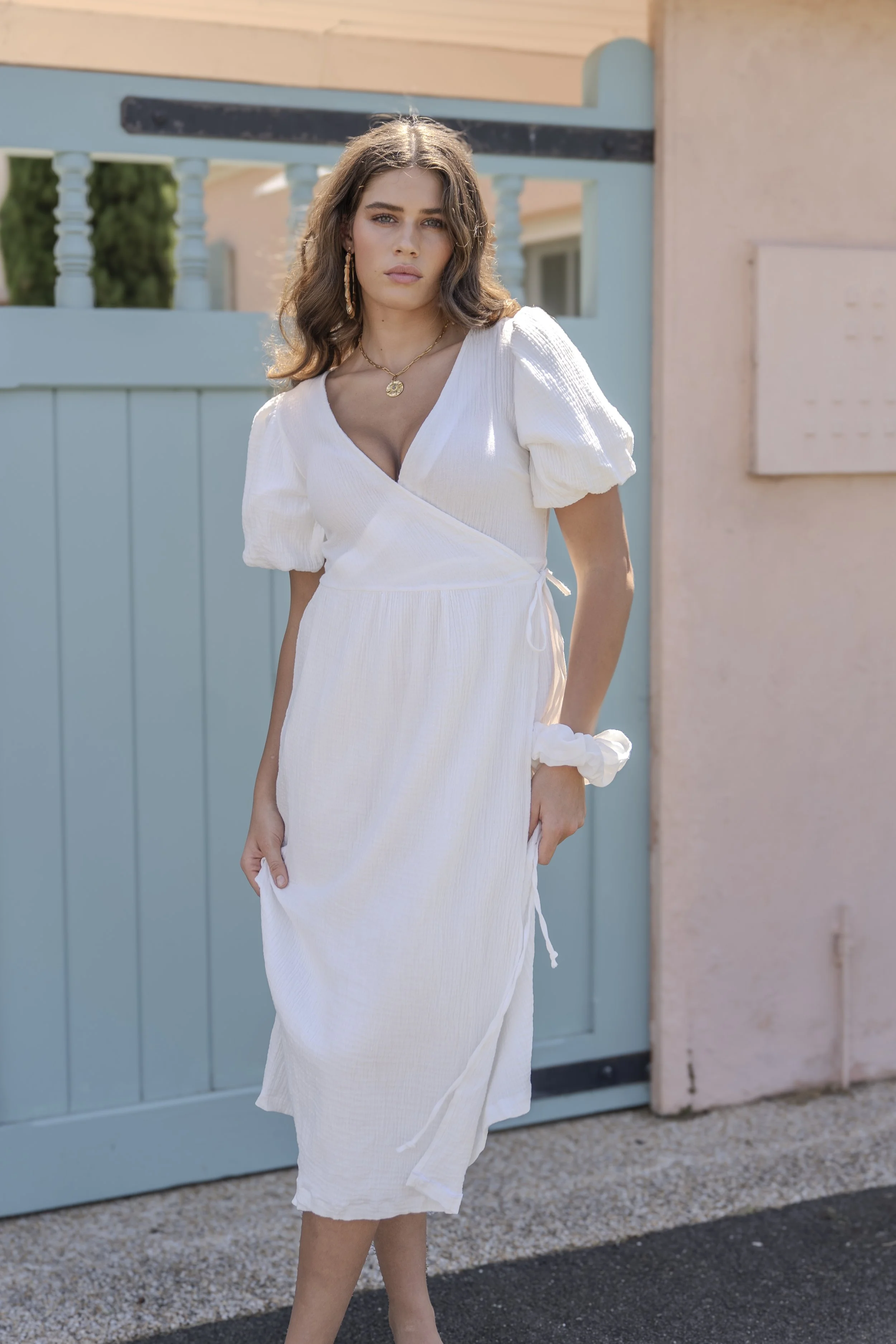 A woman with wavy brown hair wearing a white dress with puffed sleeves, standing outdoors in front of a light blue gate and pastel-colored wall.