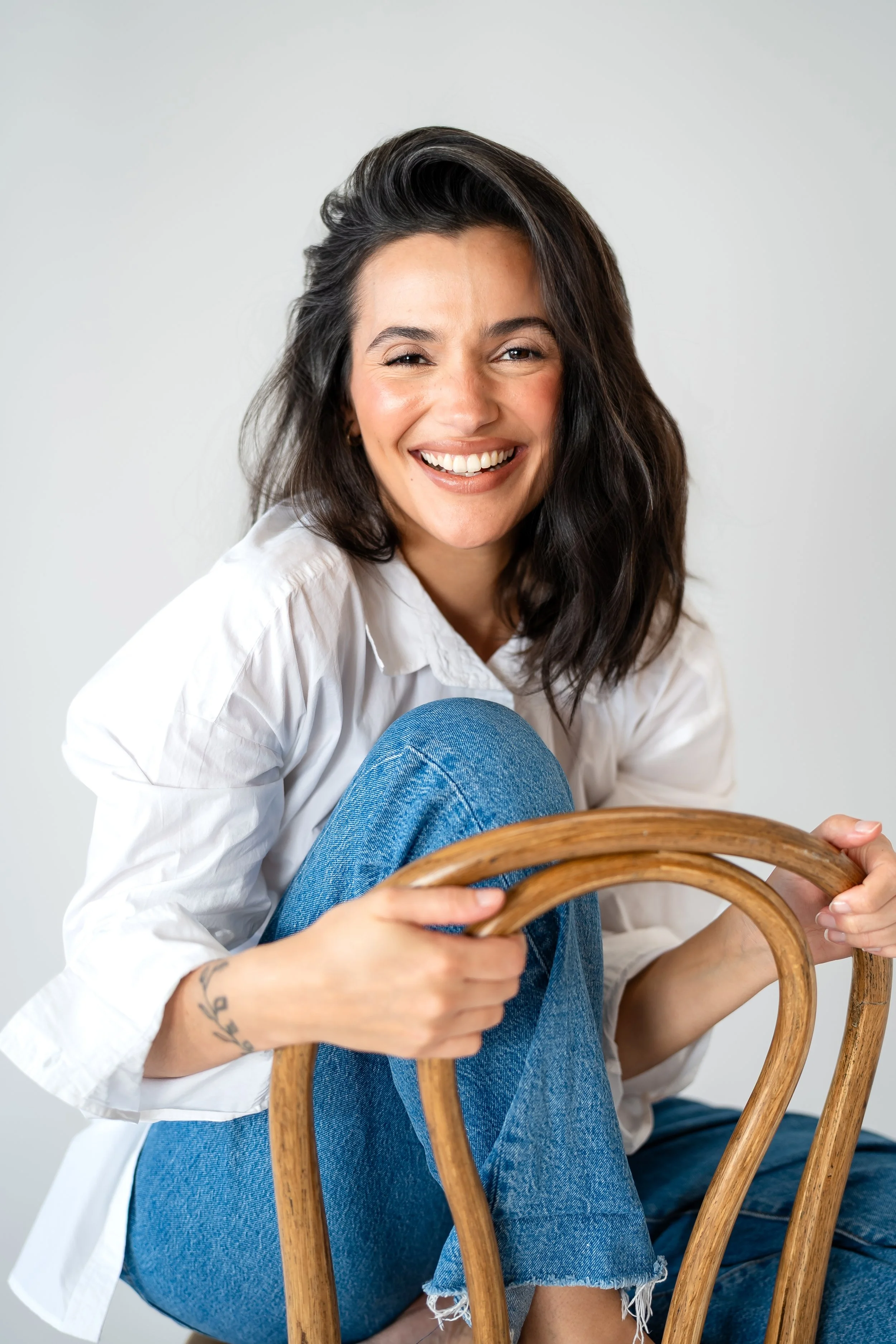 A woman with shoulder-length dark hair, smiling, wearing a white shirt and blue jeans, sitting on a wooden chair, against a plain light gray background.