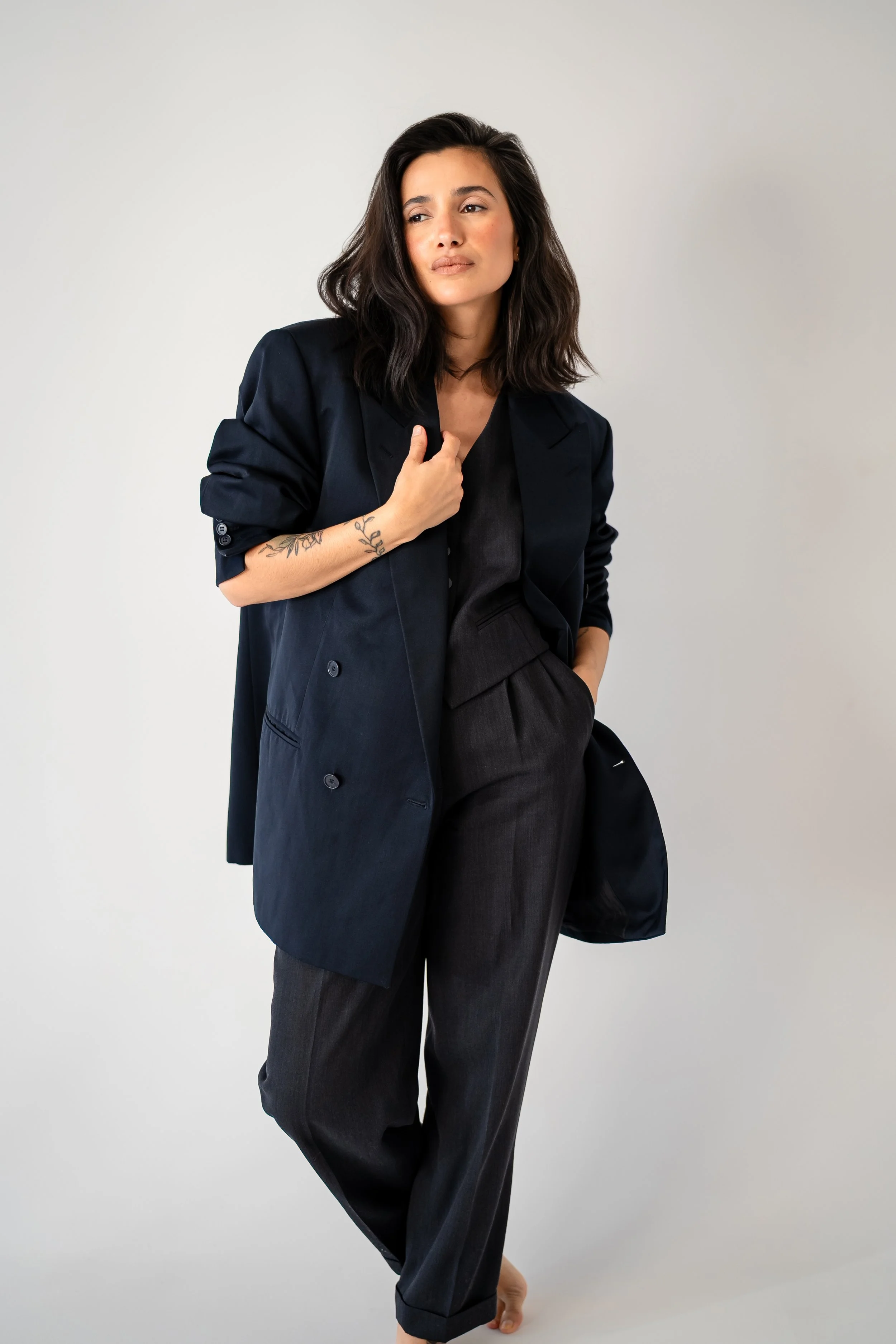 A woman with dark, wavy hair, dressed in a black suit jacket over matching pants and a black top, standing barefoot against a plain white background. She has minimal makeup and a tattoo on her left forearm.