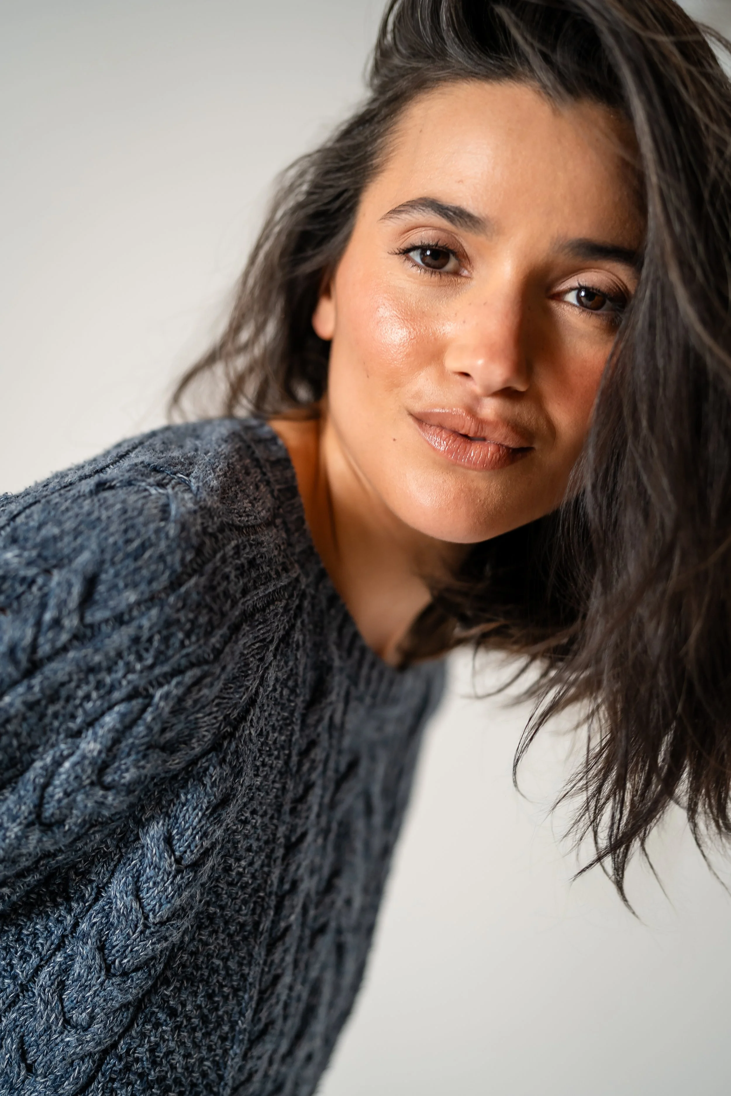 Close-up portrait of a young woman with brown eyes and dark brown wavy hair, wearing a dark knitted sweater, looking at the camera with a subtle smile.