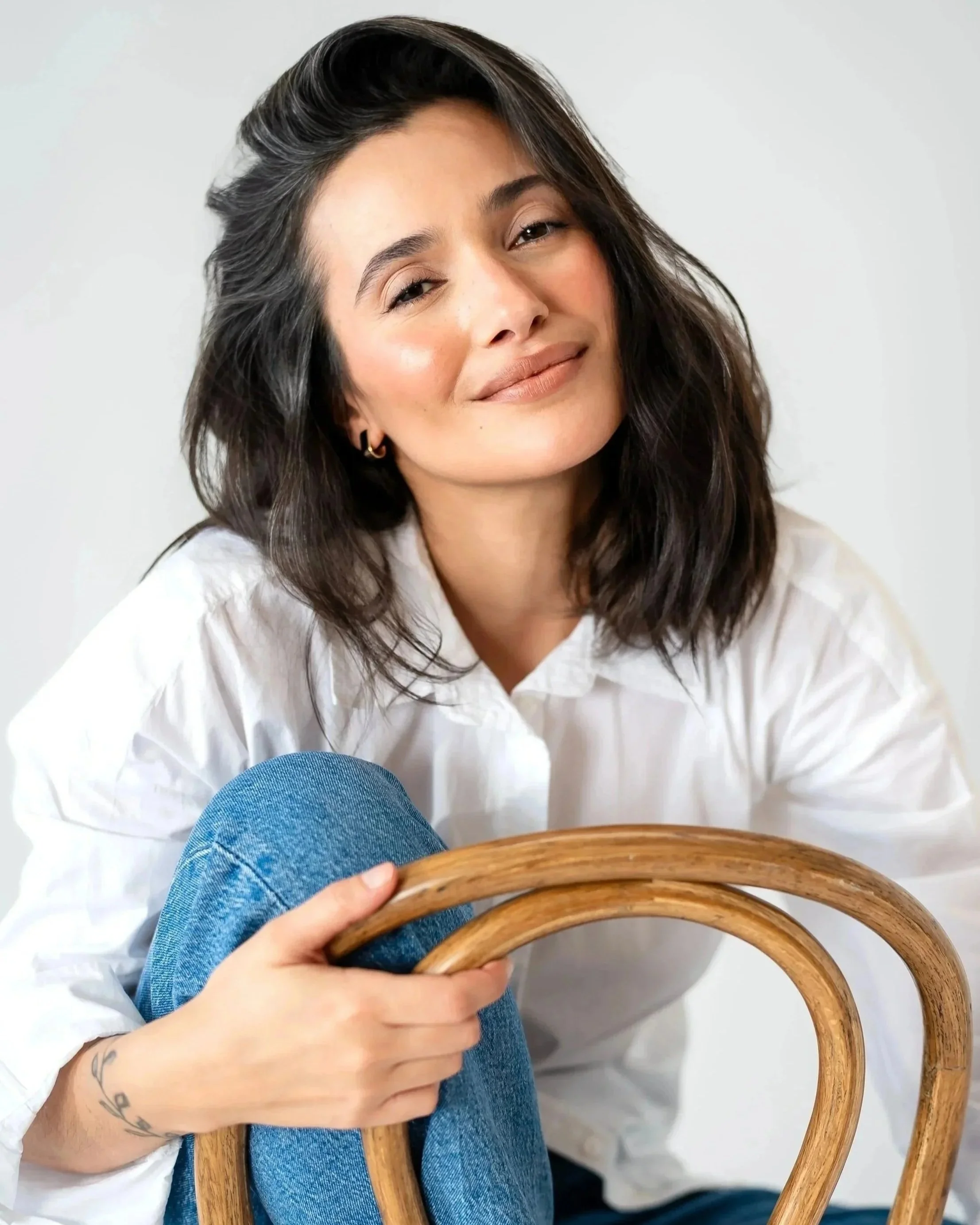 A woman with shoulder-length dark hair, wearing a white shirt, sitting on a wooden chair, smiling softly, with her head slightly tilted. She holds the back of the chair with her hand, which displays a small tattoo.