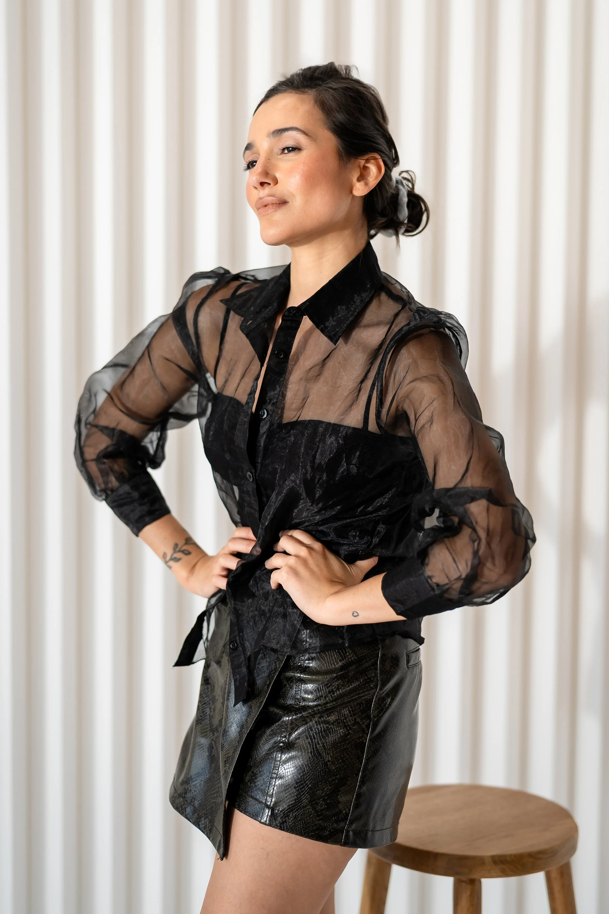 A woman with dark hair tied back, wearing a sheer black blouse with puffed sleeves and black leather shorts, standing with hands on hips in front of cream-colored curtains.