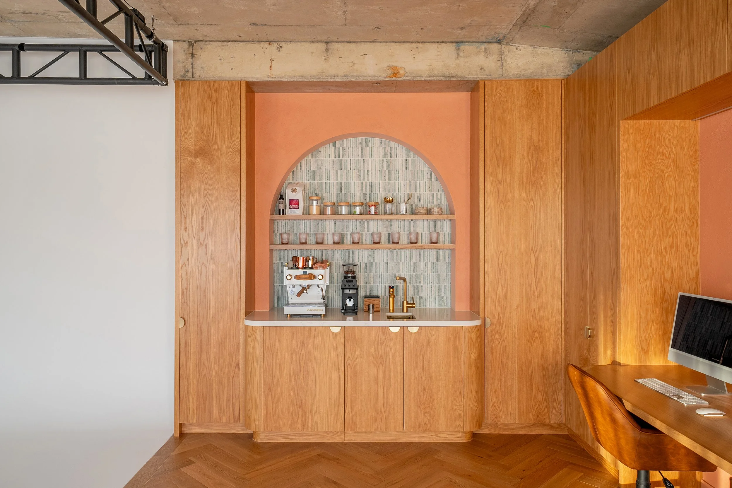 A modern kitchen nook with a wooden cabinet background, a small countertop, shelves with jars and cups, a white espresso machine, a coffee grinder, a gold faucet, and a computer on a wooden desk.