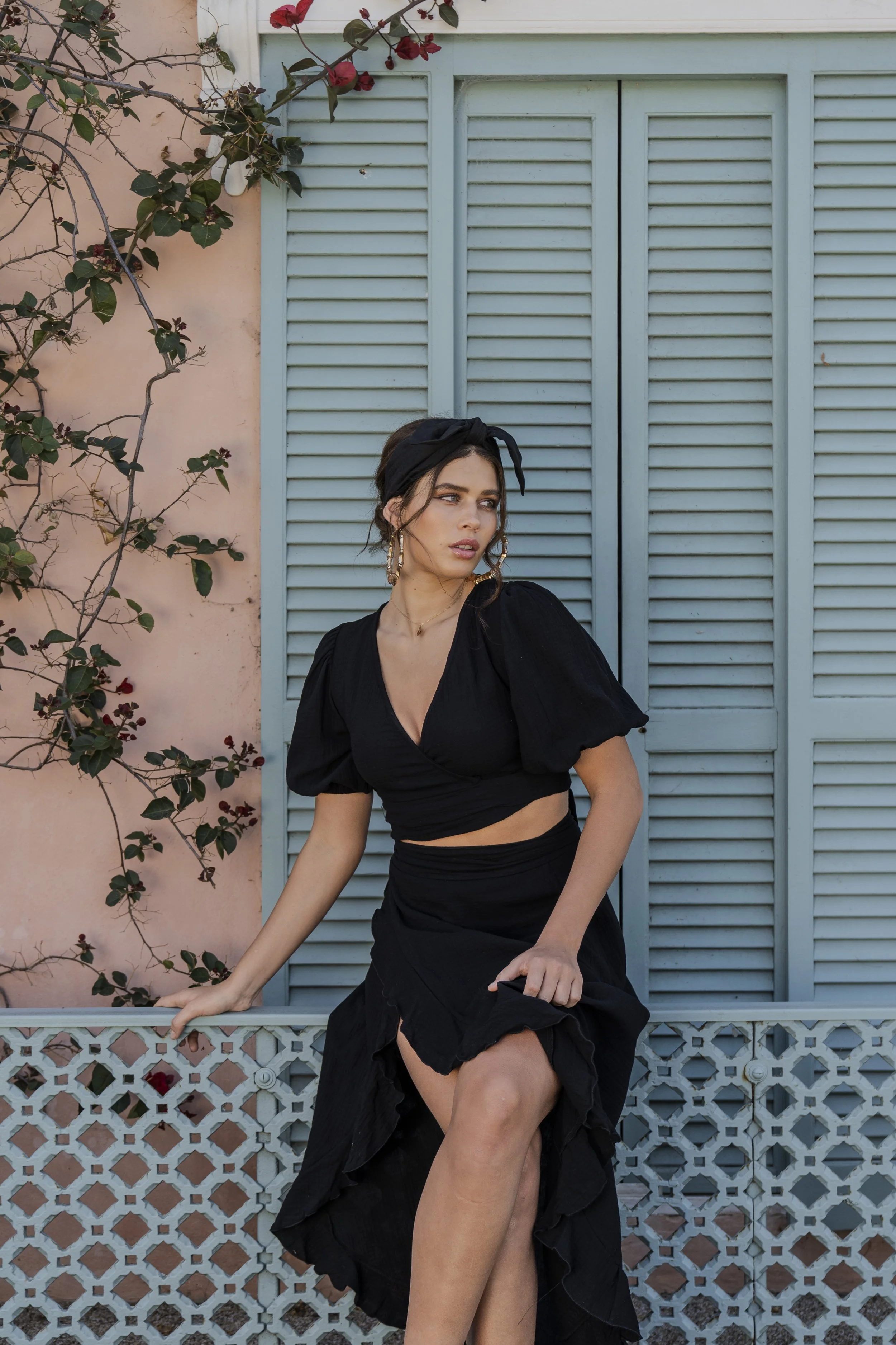 A woman wearing a black outfit with a crop top and long skirt with a slit, posing indoors against a backdrop of pale blue shutters and a peach-colored wall with climbing plants and red flowers.