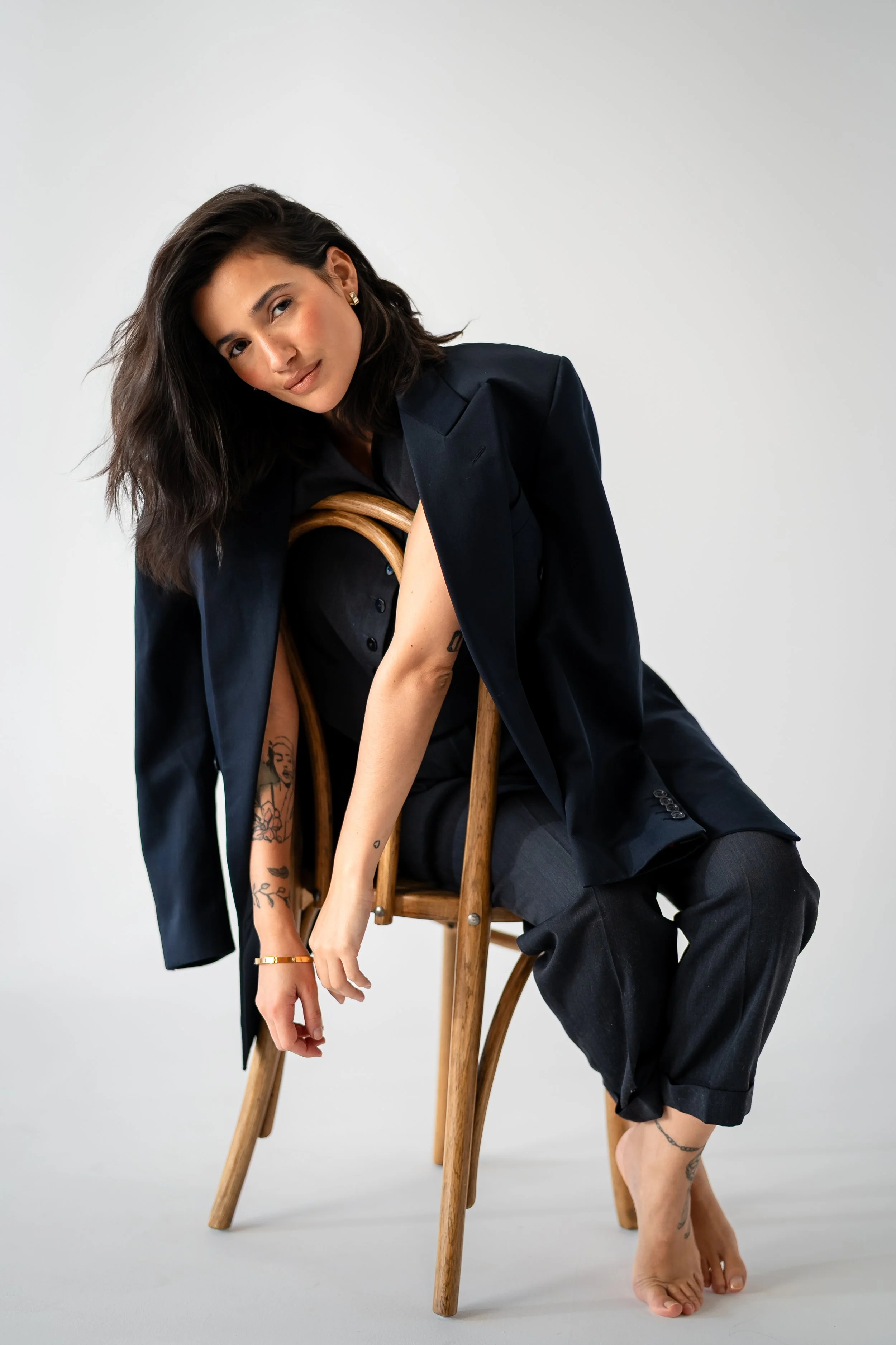 Woman with dark hair, tattoos, and light skin sitting on a wooden chair, wearing a black blazer, black dress, and gray pants, barefoot, against a plain white background.
