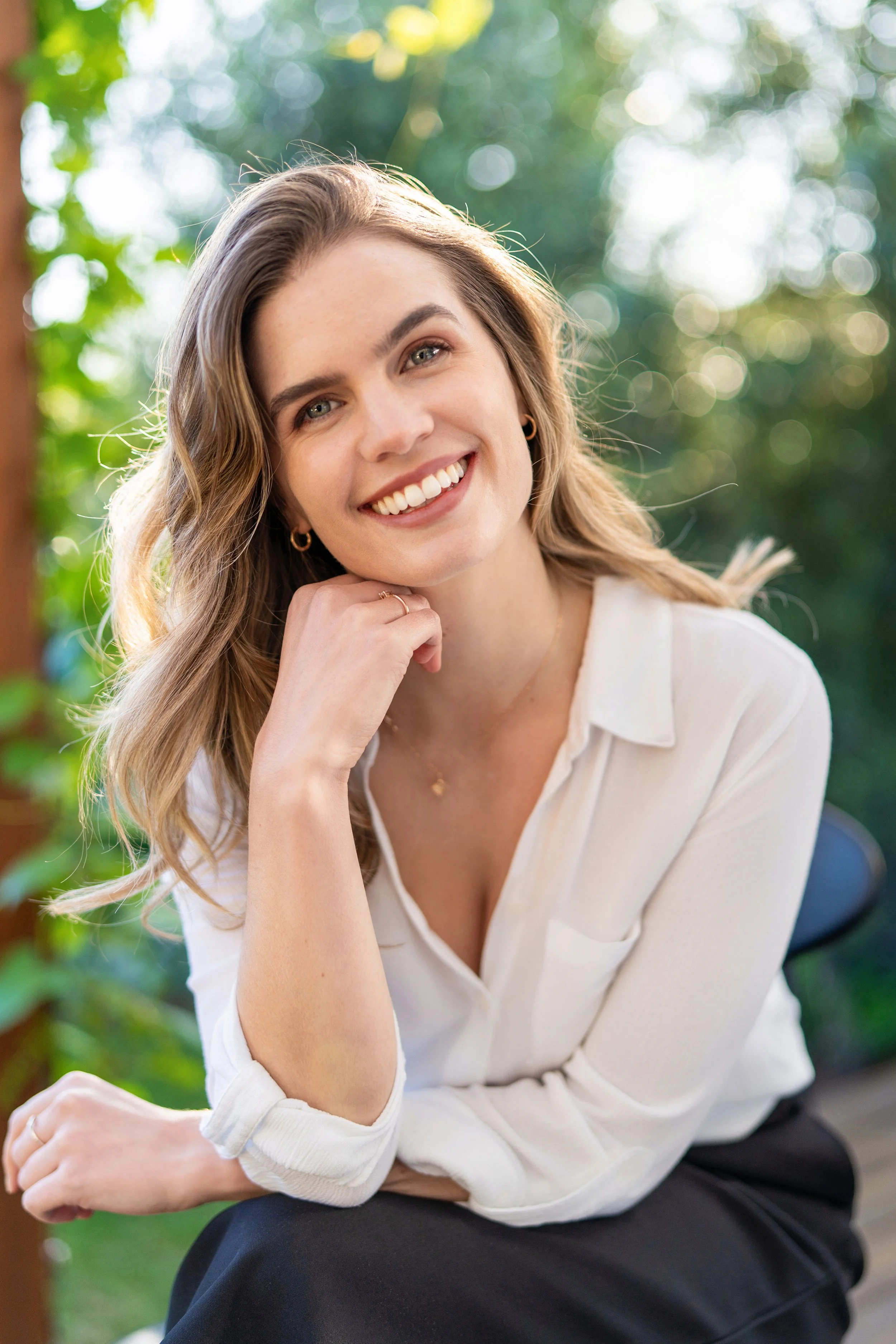 Smiling woman with wavy hair wearing a white blouse and black skirt, sitting outdoors with blurred greenery in the background.