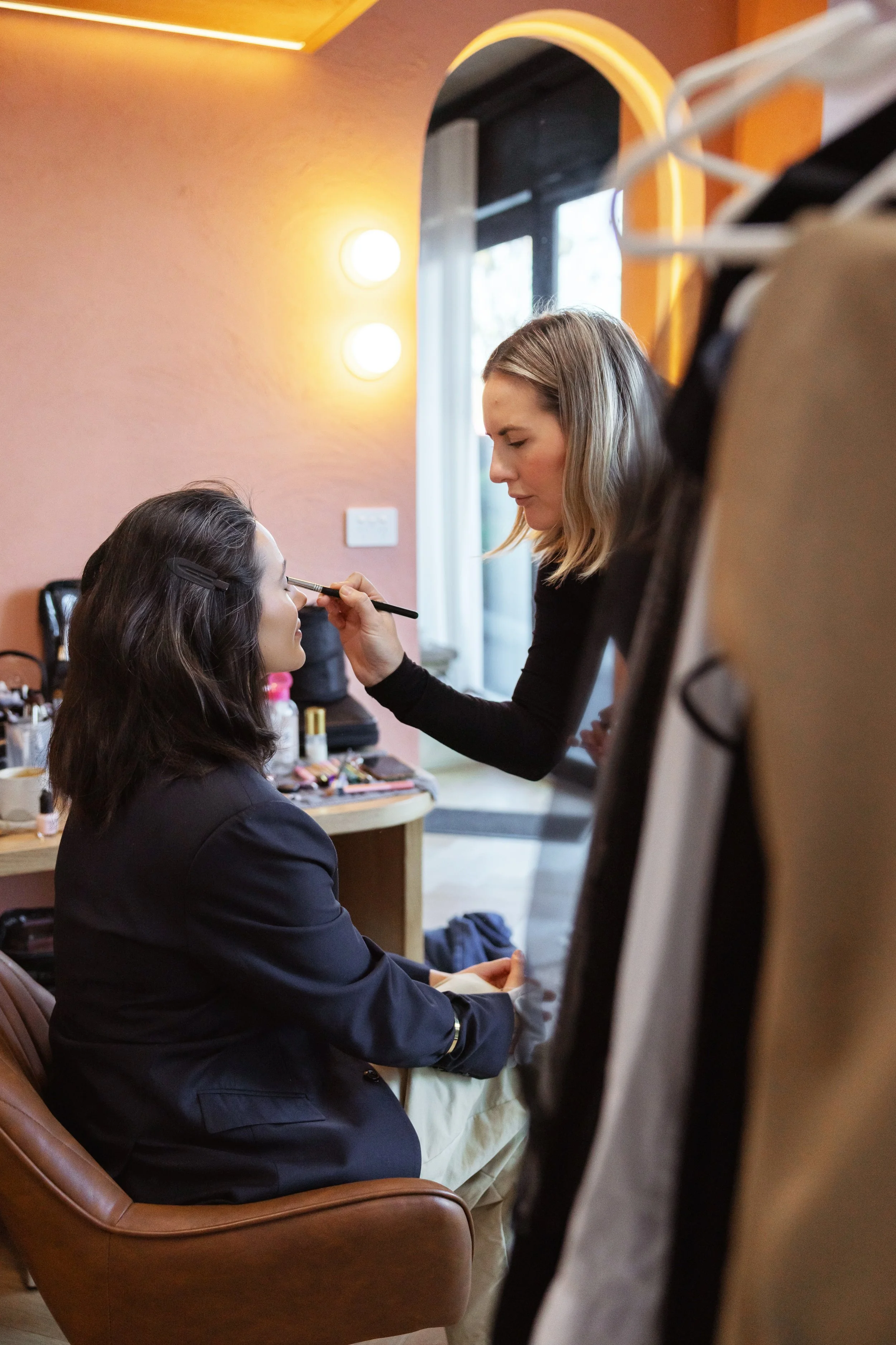 A woman is applying makeup to another woman seated in a chair in a room with warm lighting.
