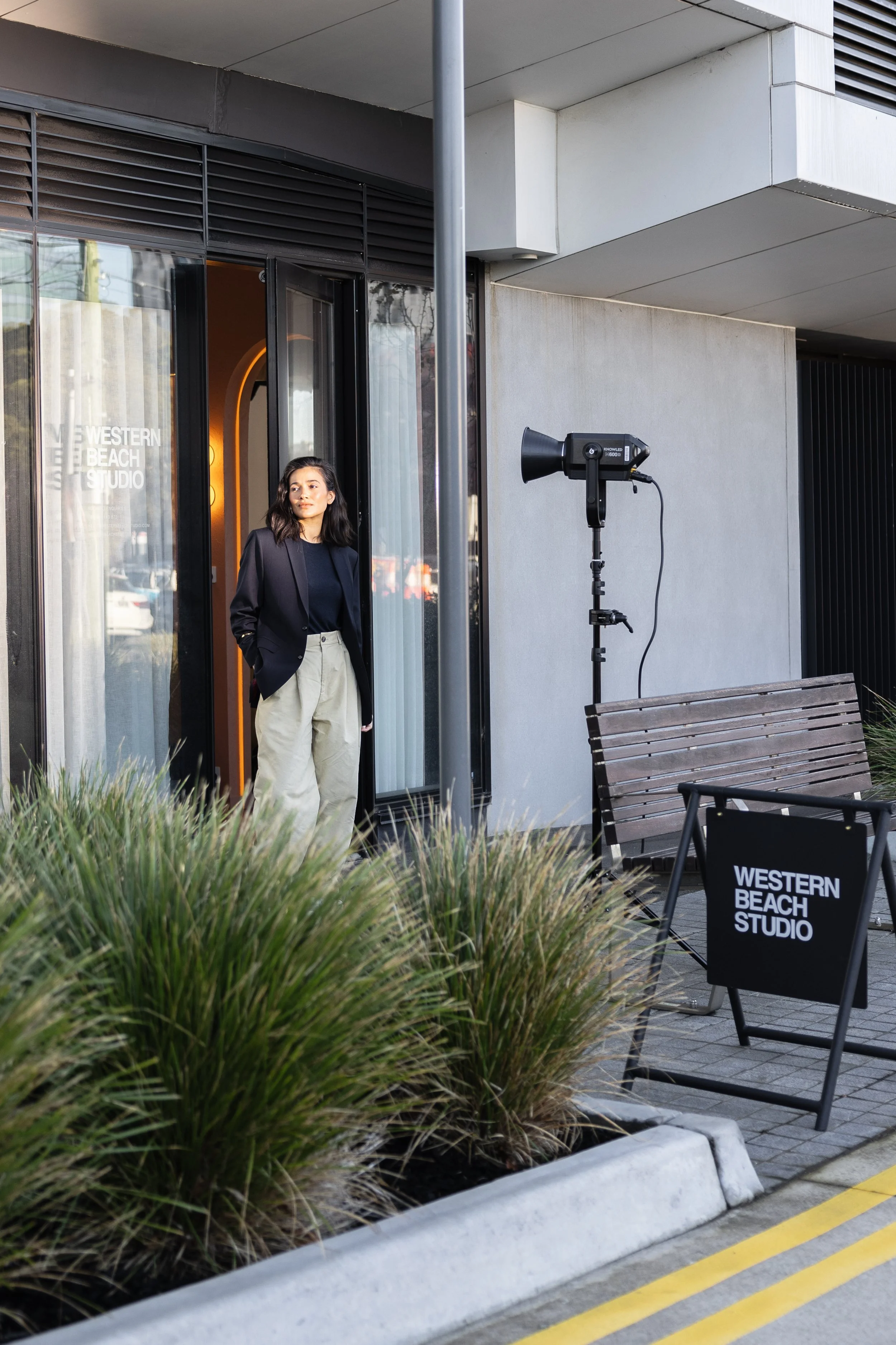 A woman standing outside the Western Beach Studio, near a photography setup with a large studio light, a wooden bench, and a sign that reads Western Beach Studio. She is wearing a dark blazer and light-colored pants.