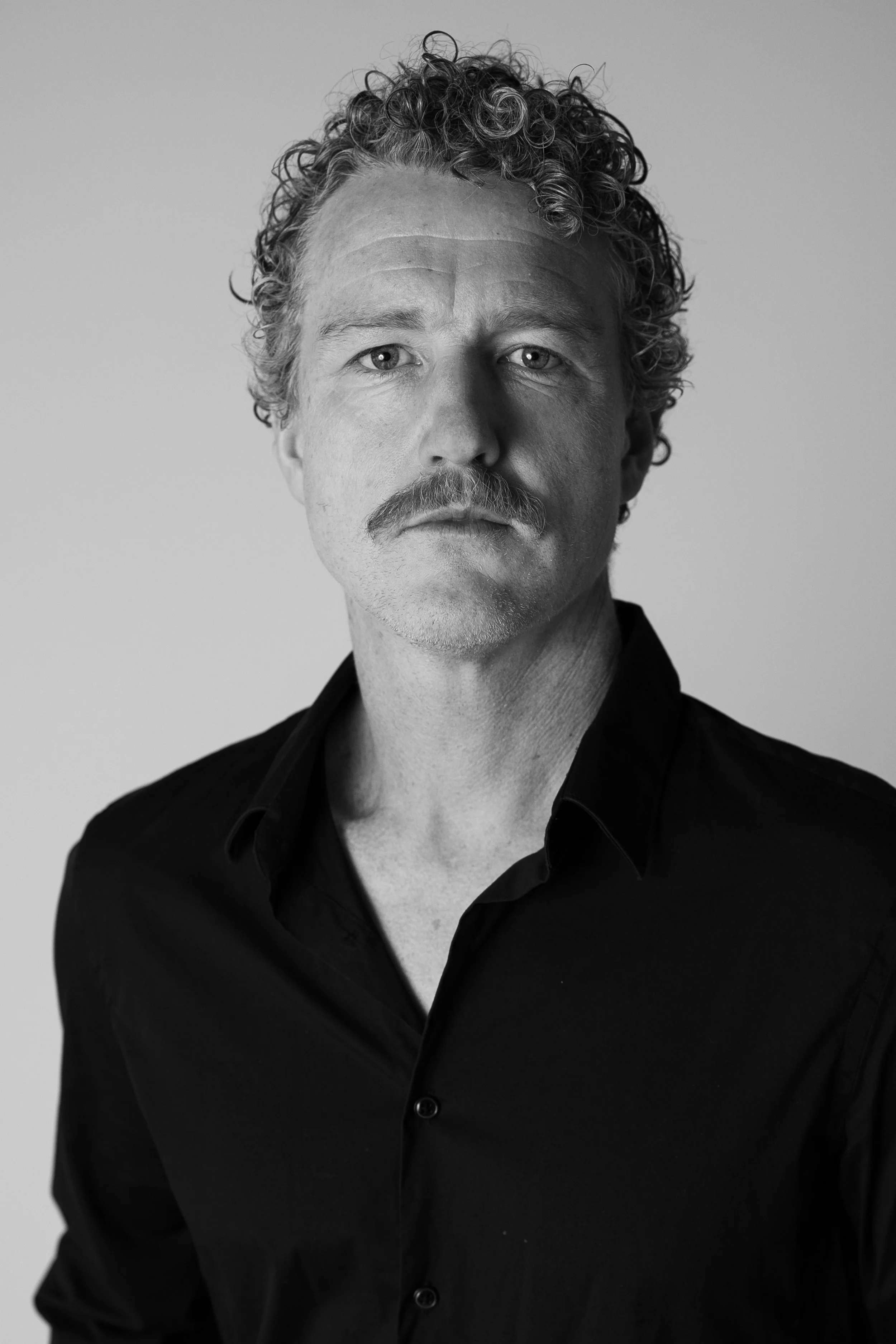 Black and white portrait of a man with curly hair and a mustache, wearing a black shirt, looking directly at the camera.