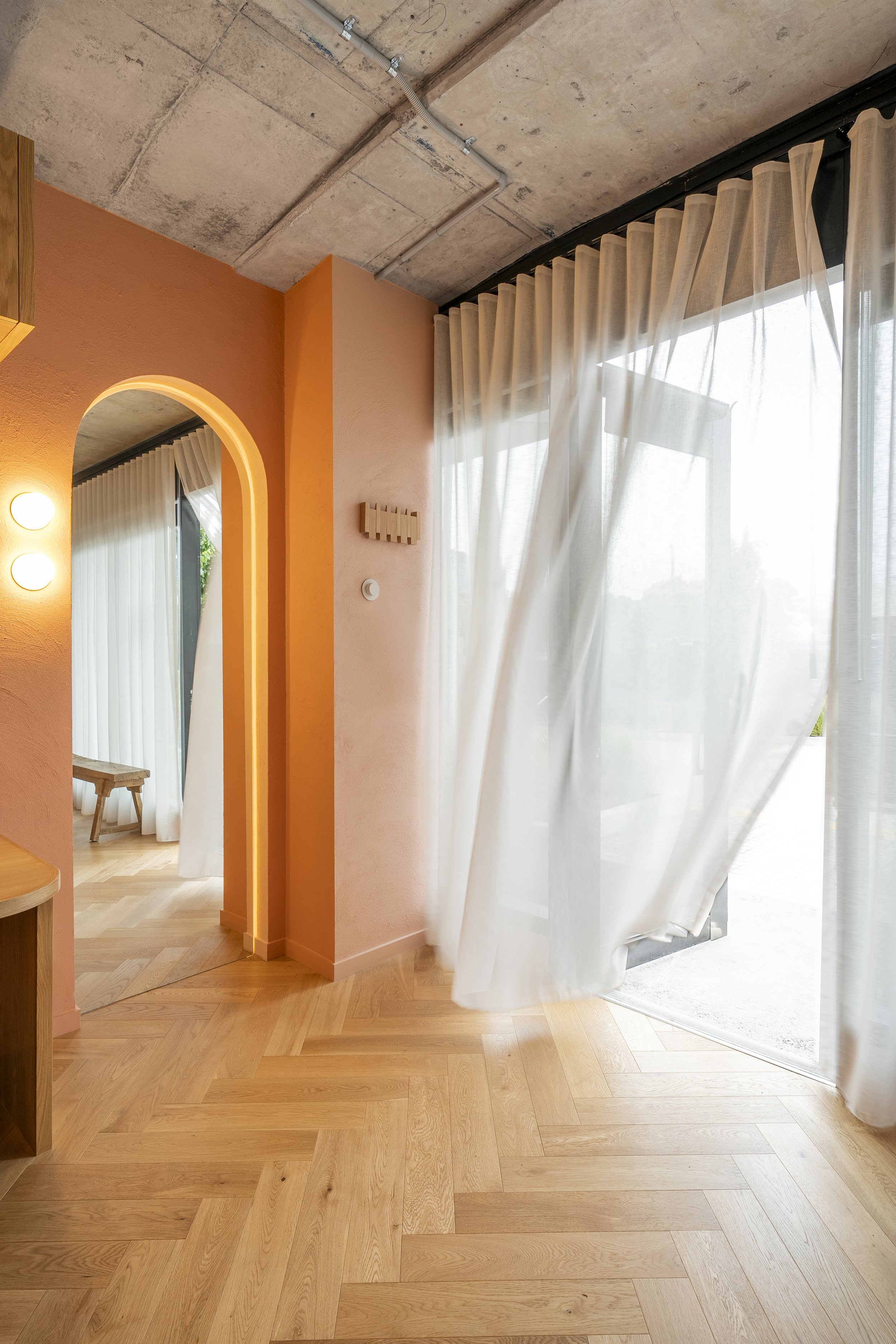 Bright room with wooden flooring, peach-colored wall, and large window with sheer white curtains. Part of an outdoor space is visible through the window.
