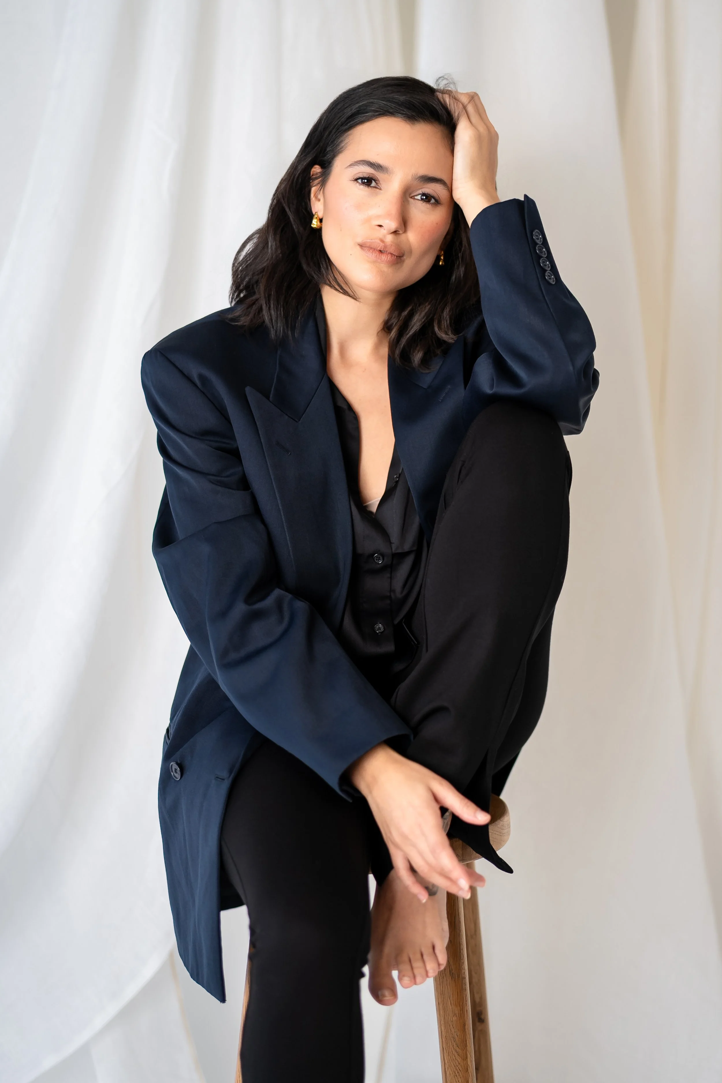 A woman with dark hair wearing a navy blazer and black pants sitting on a wooden stool, with a neutral background.