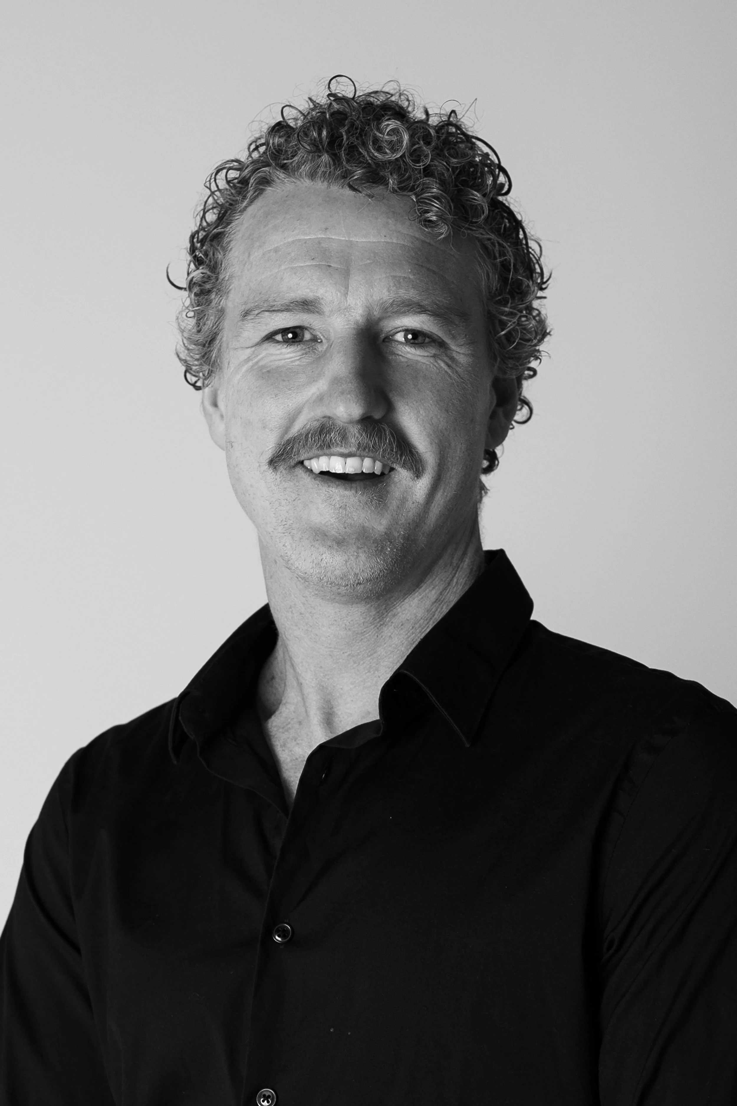 Black and white portrait of a smiling man with curly hair, mustache, and wearing a dark shirt.