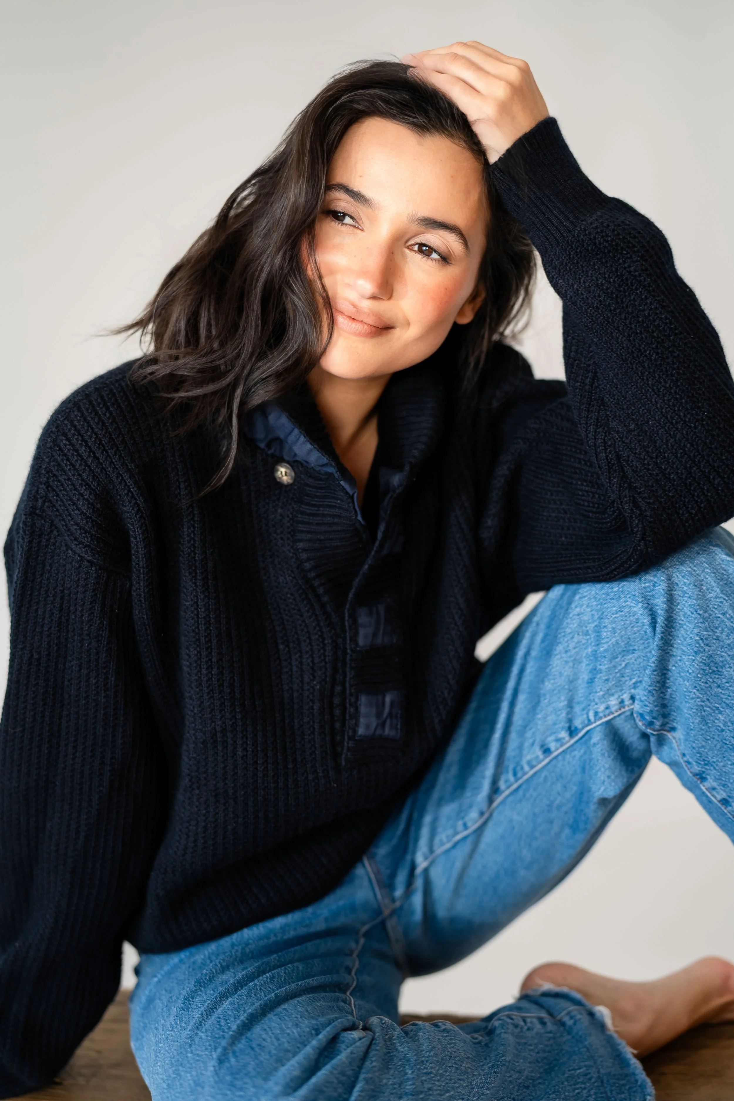 A young woman with dark wavy hair sitting on the floor, wearing a black sweater and blue jeans, with one hand on her head and a slight smile.