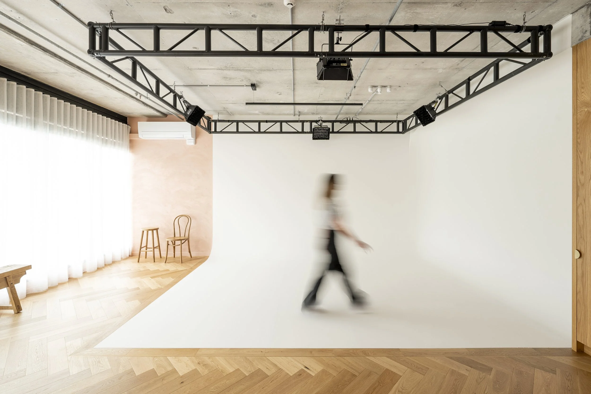 A person walking in a photography studio with a white backdrop and wooden flooring; studio equipment and chairs visible in the background.
