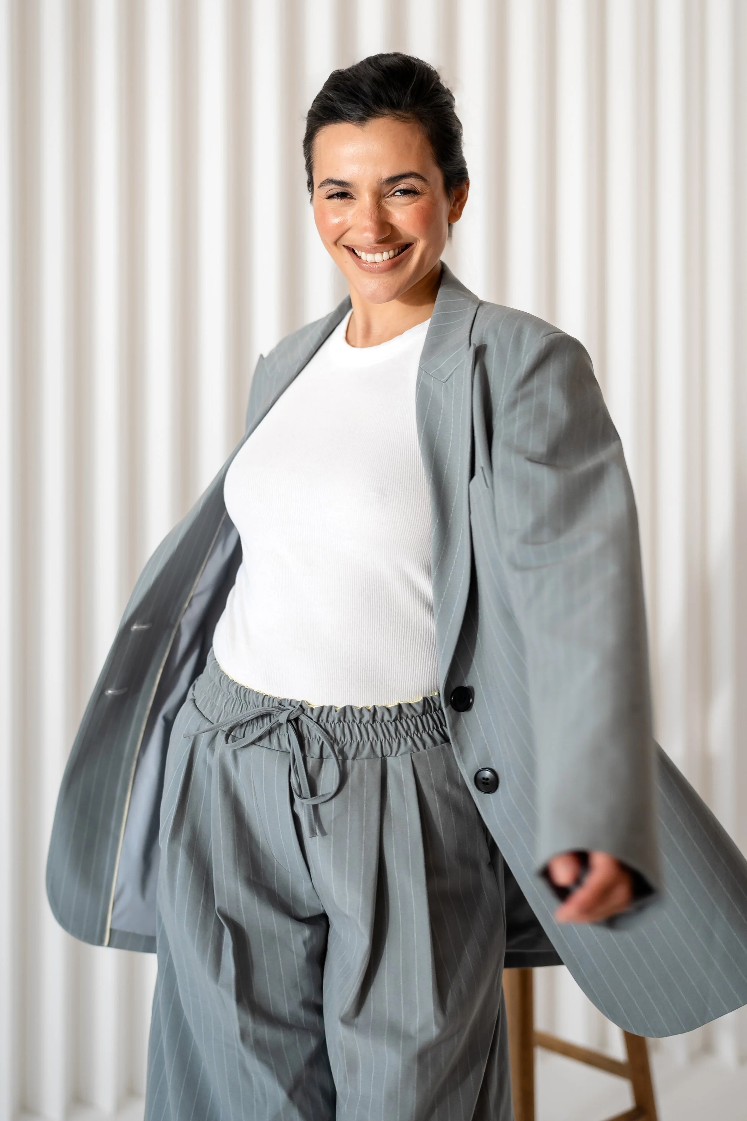A woman with dark hair styled back, wearing a light gray pinstripe suit with a matching blazer and wide-leg pants, over a white t-shirt, smiling and holding the edge of her blazer in front of a neutral curtain background.