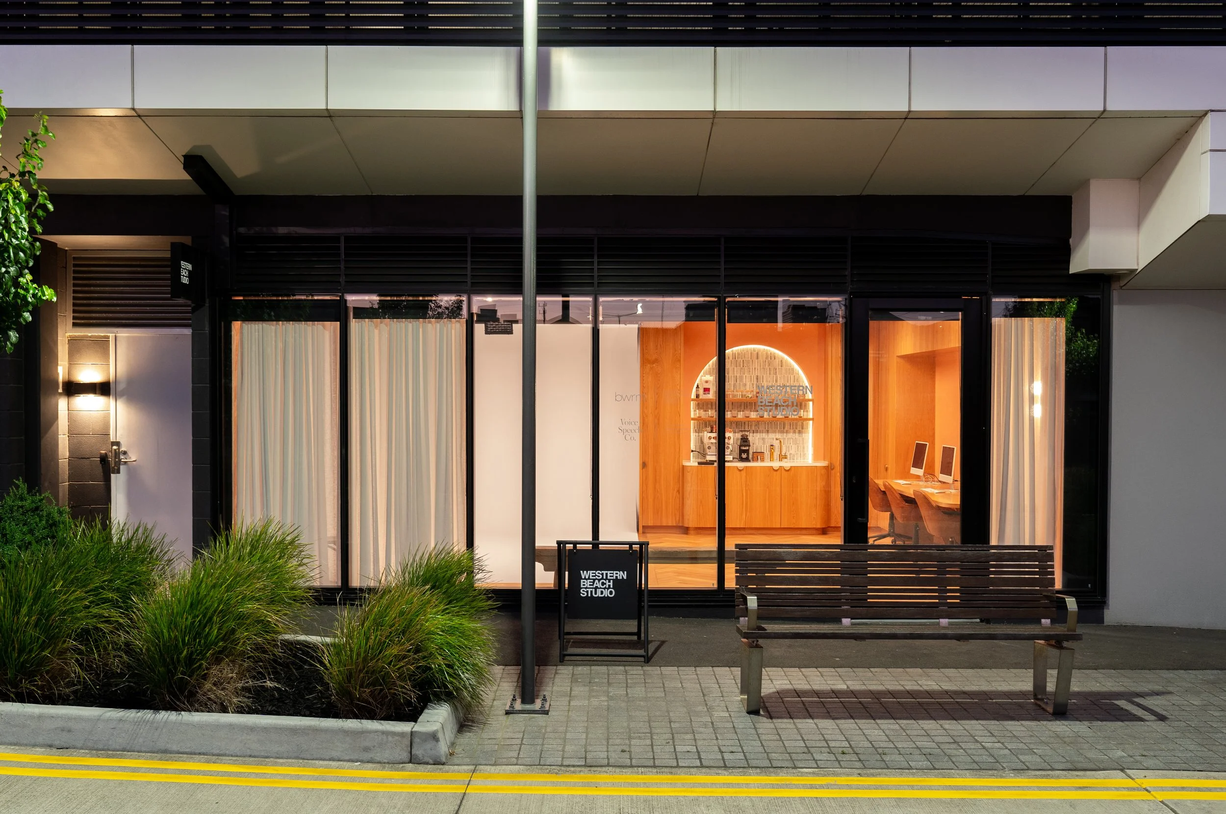 Exterior view of Western Beach Studio with large glass windows and wooden interior, a bench and a sidewalk with yellow lines.