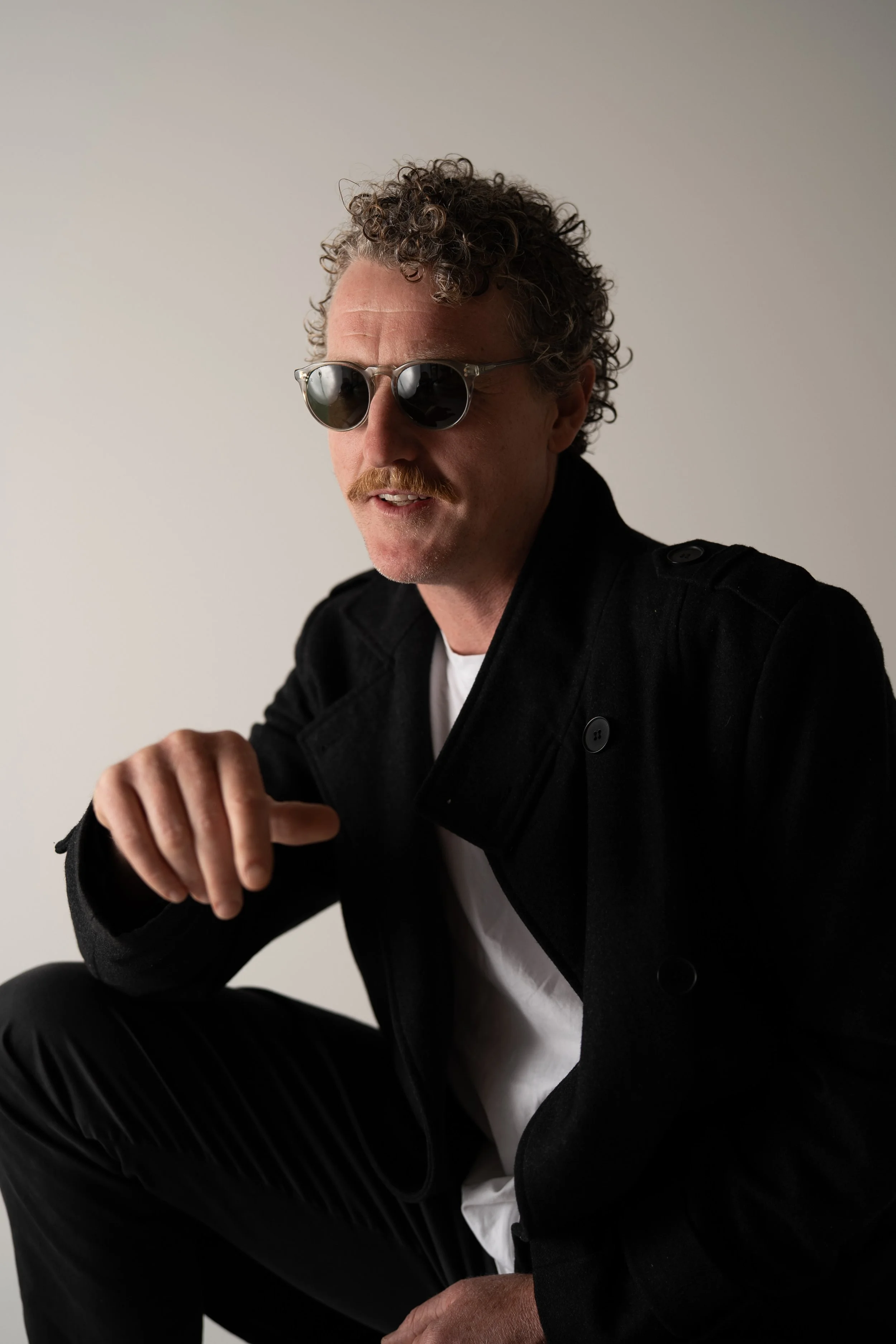 A man with curly hair, sunglasses, and a mustache wearing a black jacket, white shirt, and black pants, sitting against a plain background.