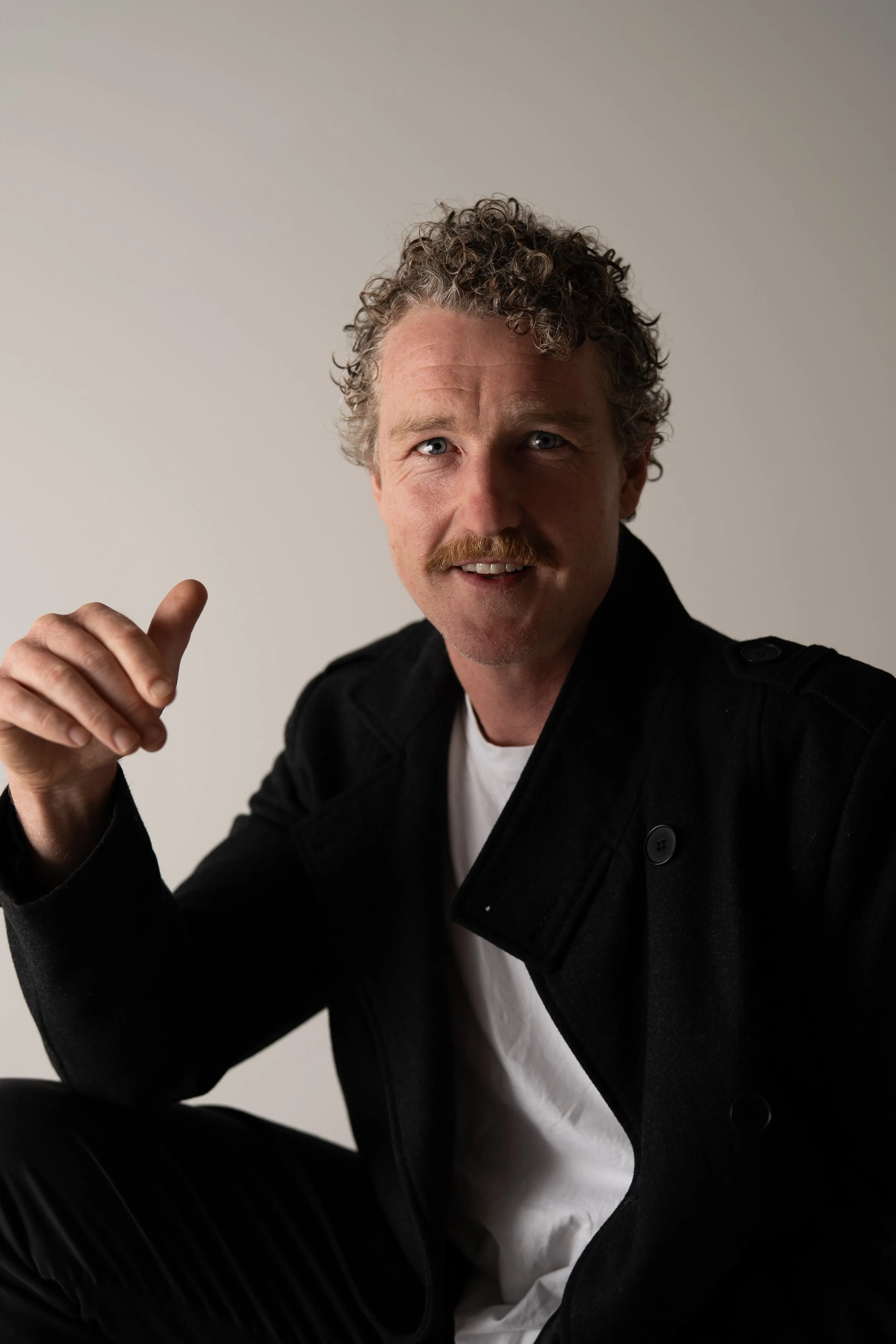 A man with curly hair and a mustache wearing a black jacket and white t-shirt, smiling and gesturing with his hand, against a plain light background.