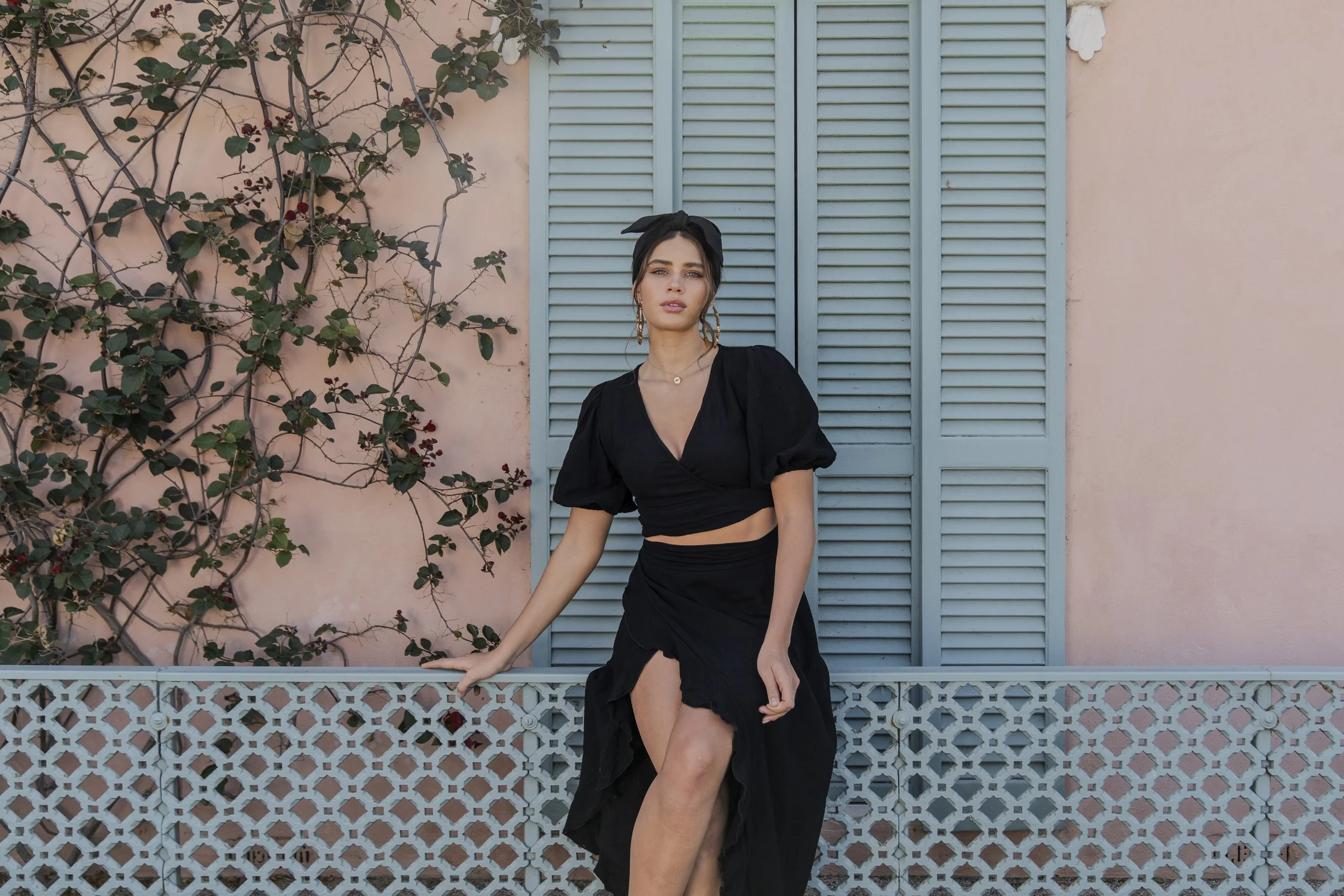 A young woman in a black dress with puffed sleeves and a slit up the leg, standing next to a decorative railing and a blue shuttered window, with a pink wall and climbing plant behind her.
