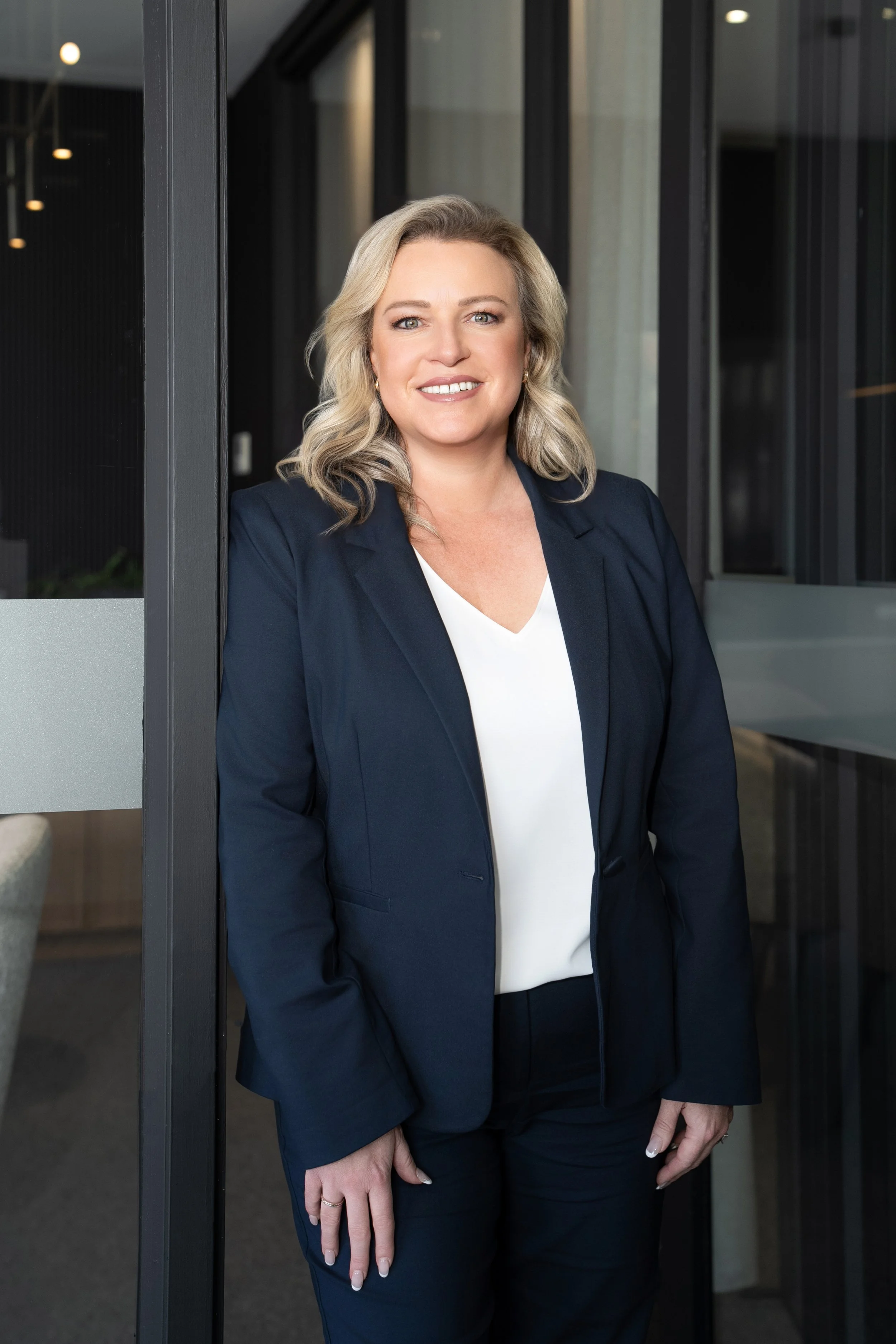 A woman with blonde hair wearing a navy blazer and white blouse standing in an office entrance.