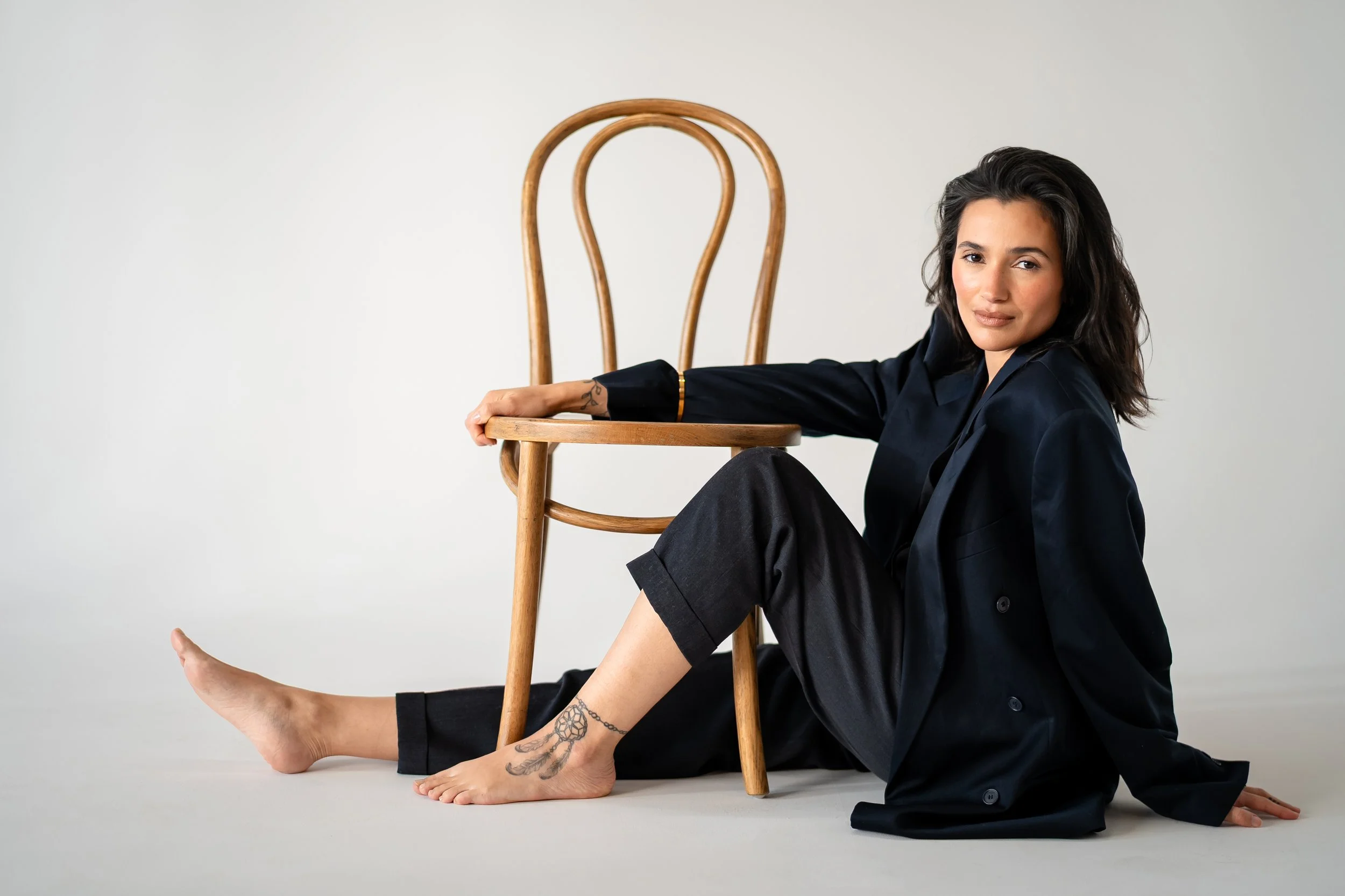A woman with dark hair, wearing a black blazer and pants, sitting on the floor with her legs extended, one arm resting on a wooden chair, and smiling at the camera. She has tattoos on her ankle and wrist, and a neutral light gray background.