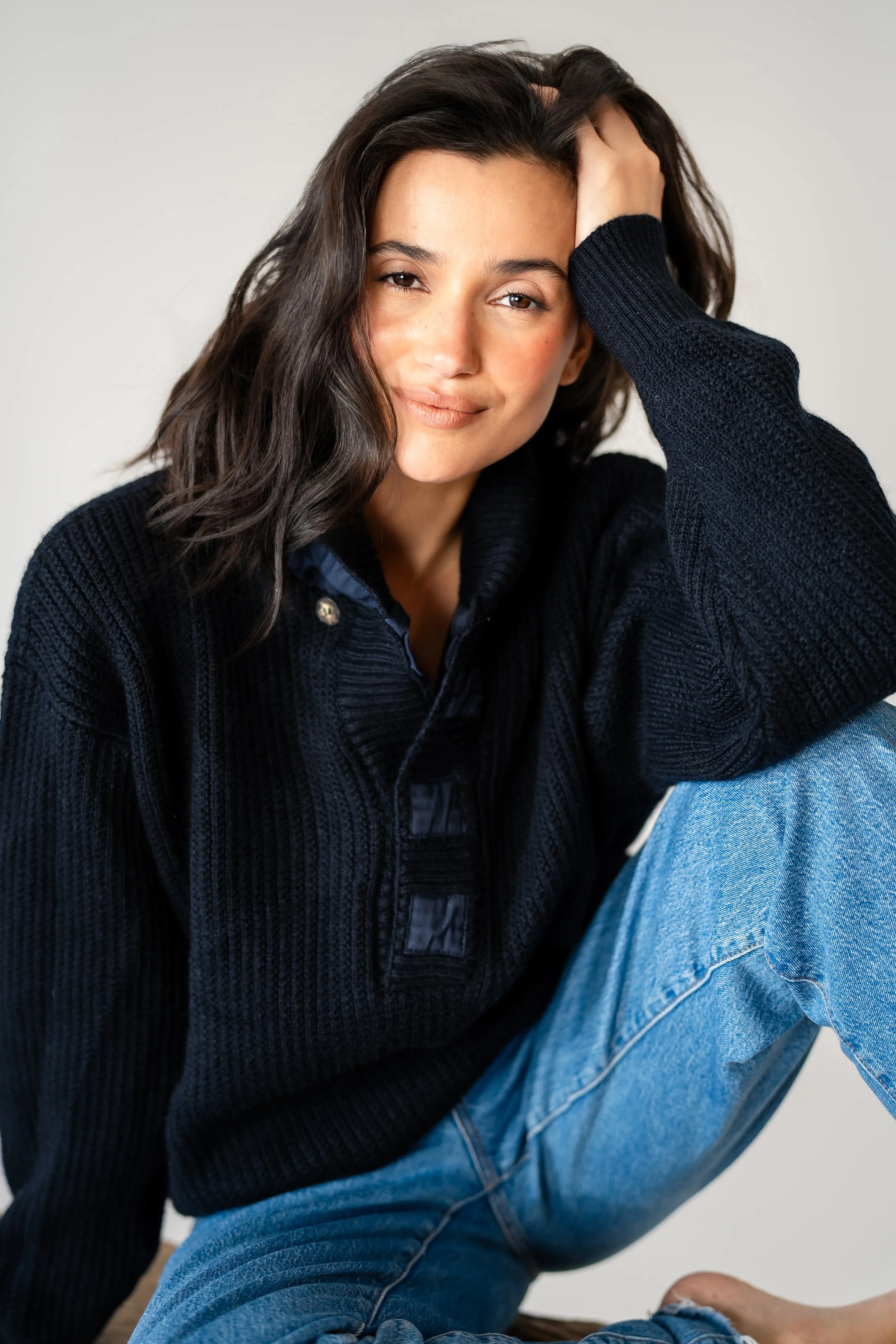 A woman with dark, wavy hair sitting against a plain background, wearing a black sweater and blue jeans, with her hand resting on her head and a relaxed, confident expression.