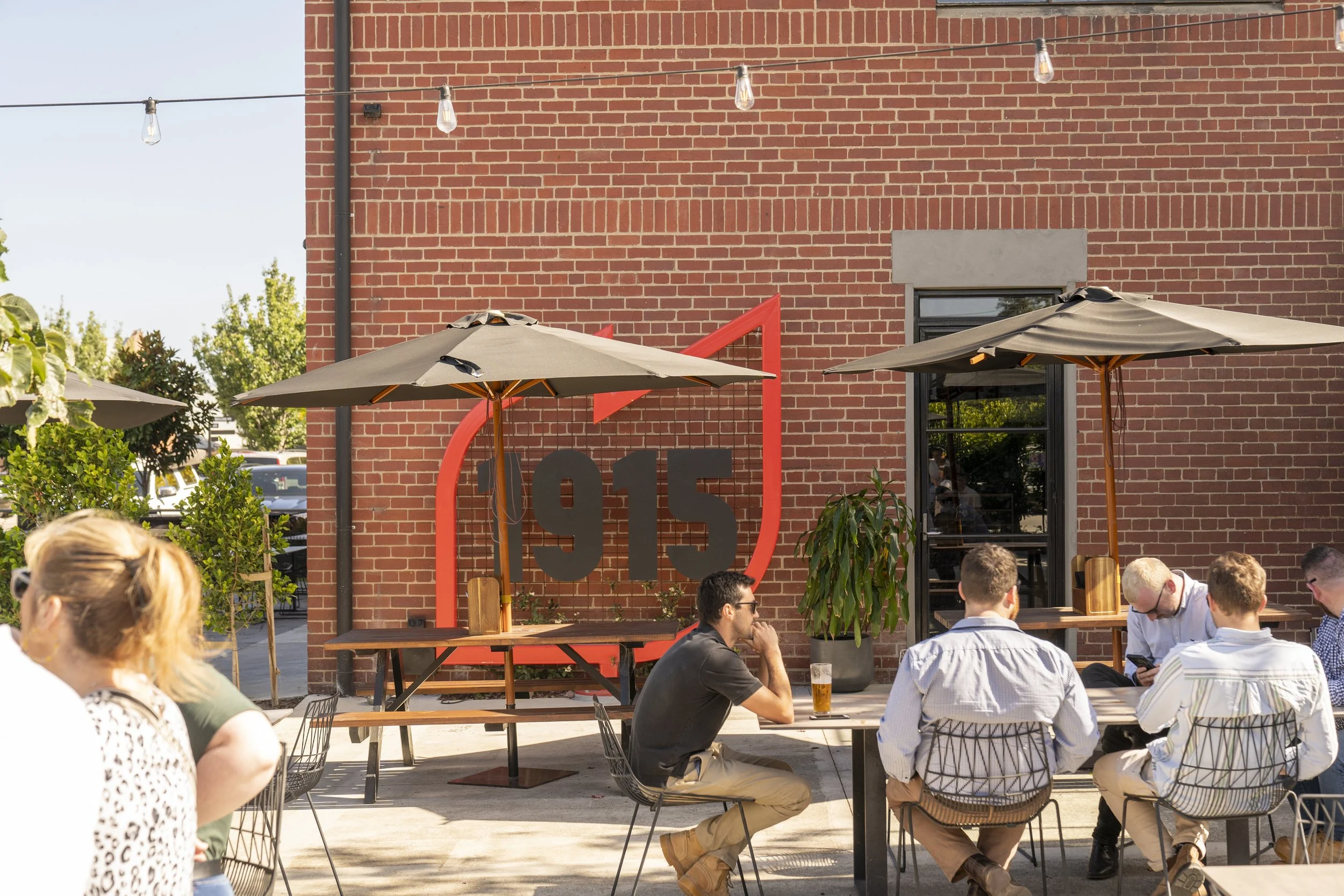 People sitting and socializing outdoors at a patio with umbrellas in front of a brick building with a '915' sign.