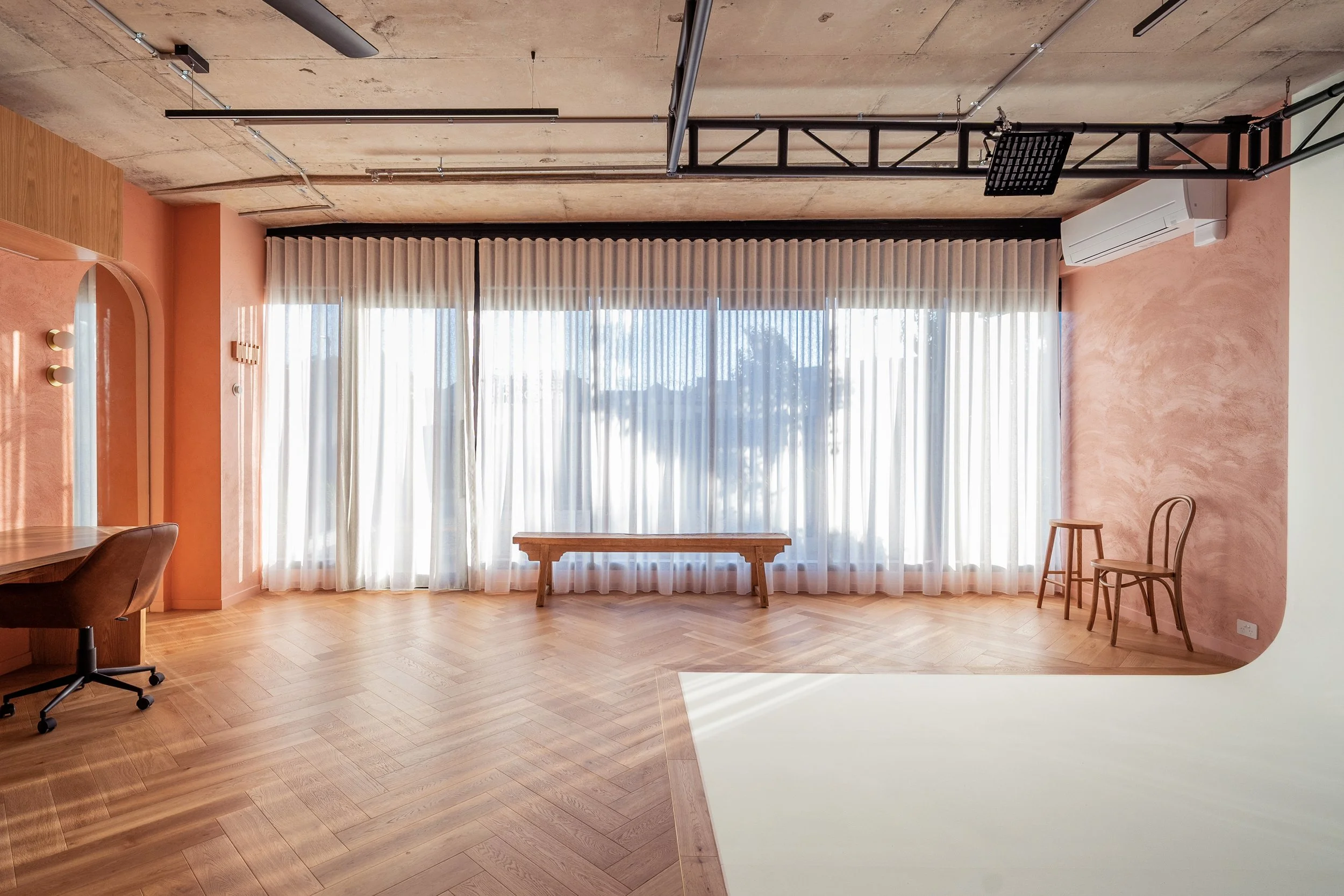 Bright living room with wooden floor, large window with white curtains, pink accent wall, simple wooden bench, two wooden chairs, desk with brown chair, ceiling with industrial-style pipes, air conditioning unit.