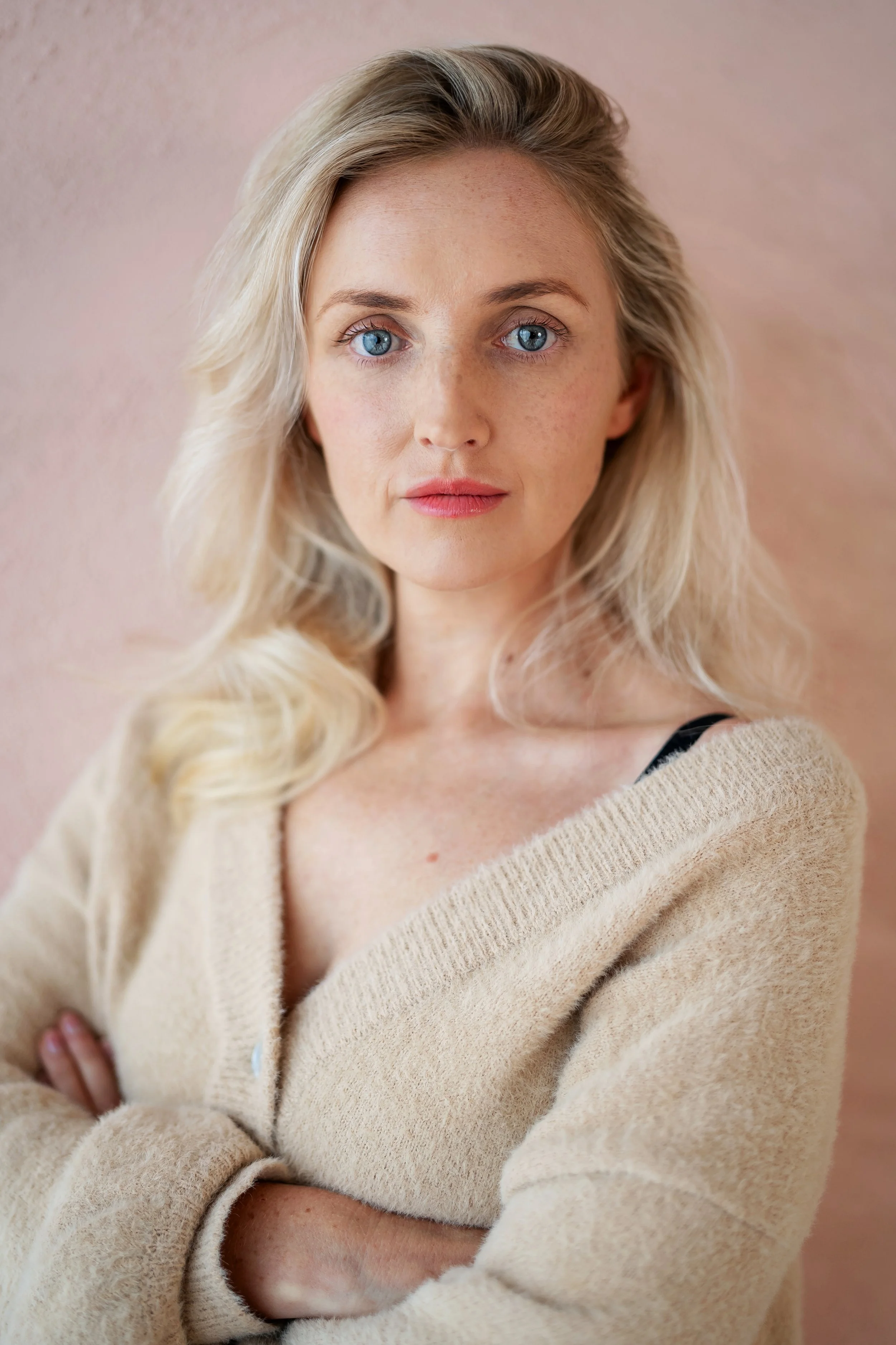 A woman with blonde hair and blue eyes wearing a beige sweater standing against a pink wall, looking directly at the camera with arms crossed.