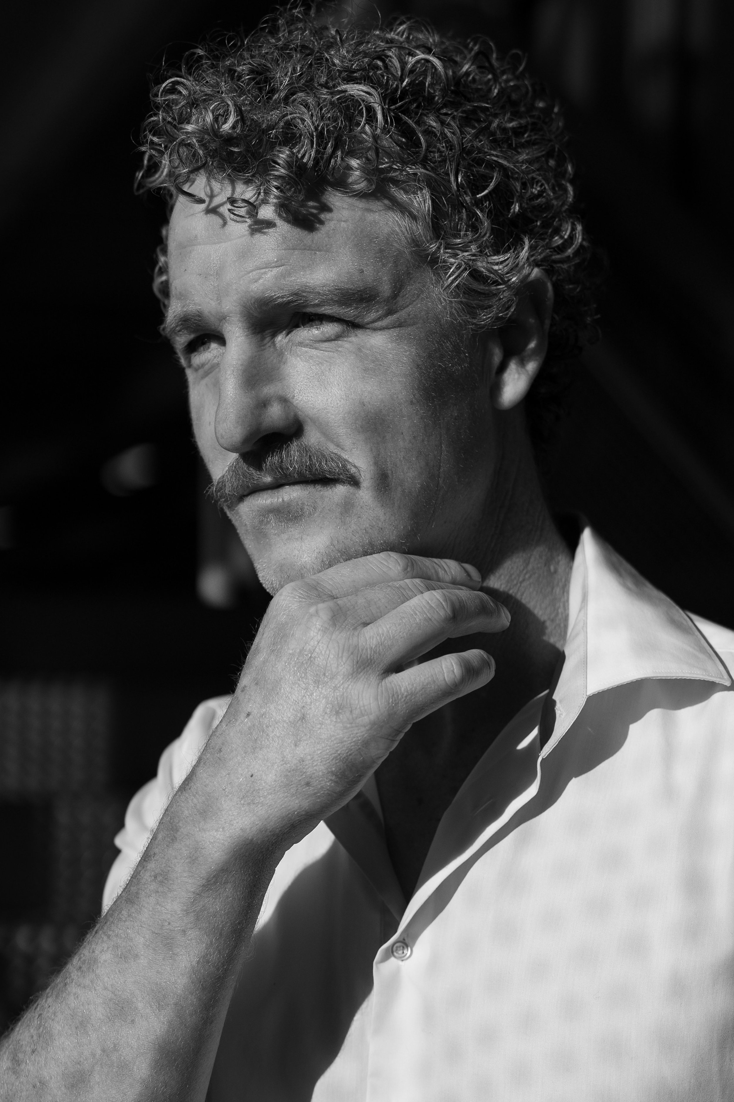 Black and white photo of a man with curly hair and a mustache, wearing a collared shirt, resting his chin on his hand.