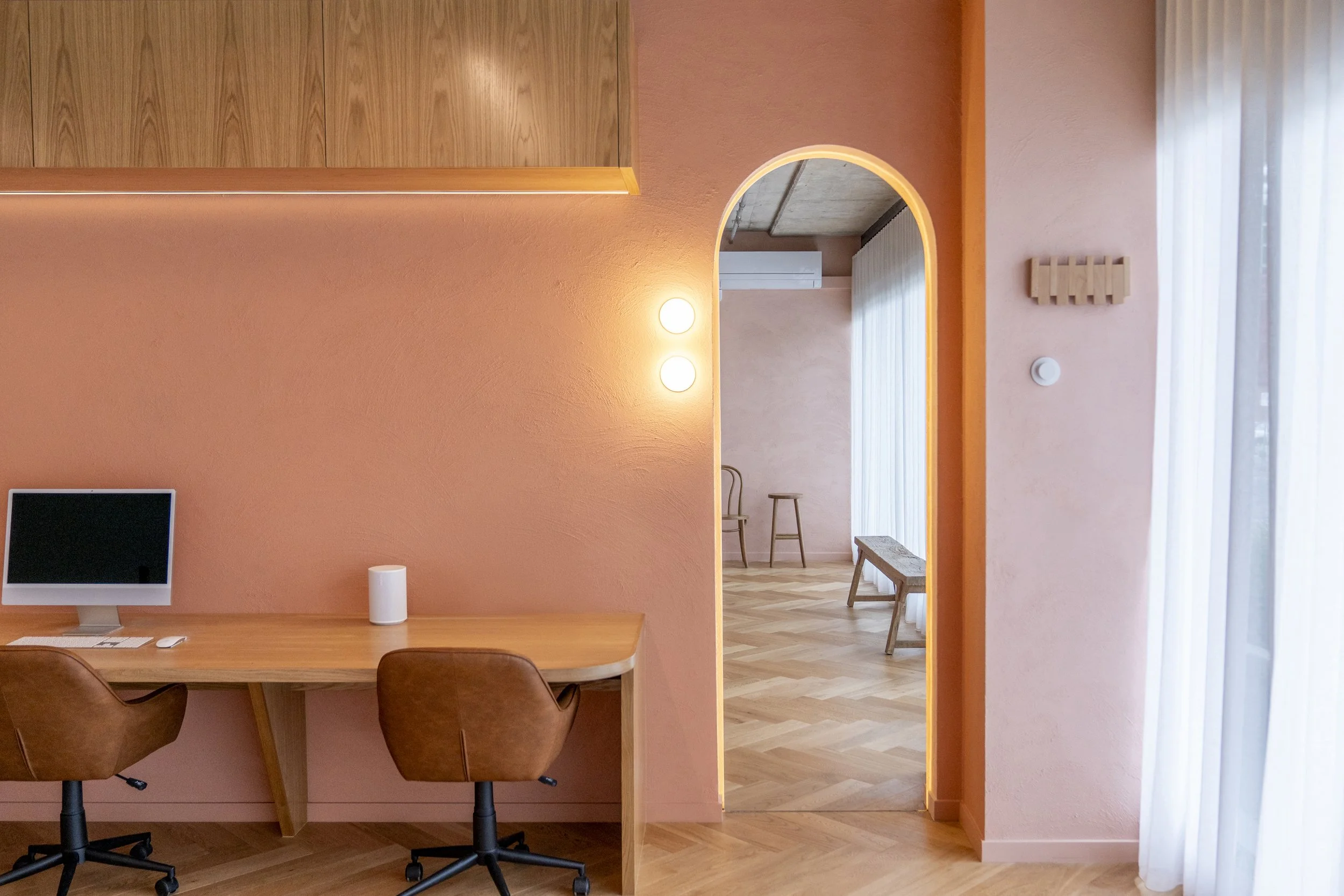 A minimalist home office with a wooden desk, a computer, two brown chairs, peach-colored walls, a doorway leading to a room with a bench and chairs, natural light from a window with white curtains, and wooden flooring.
