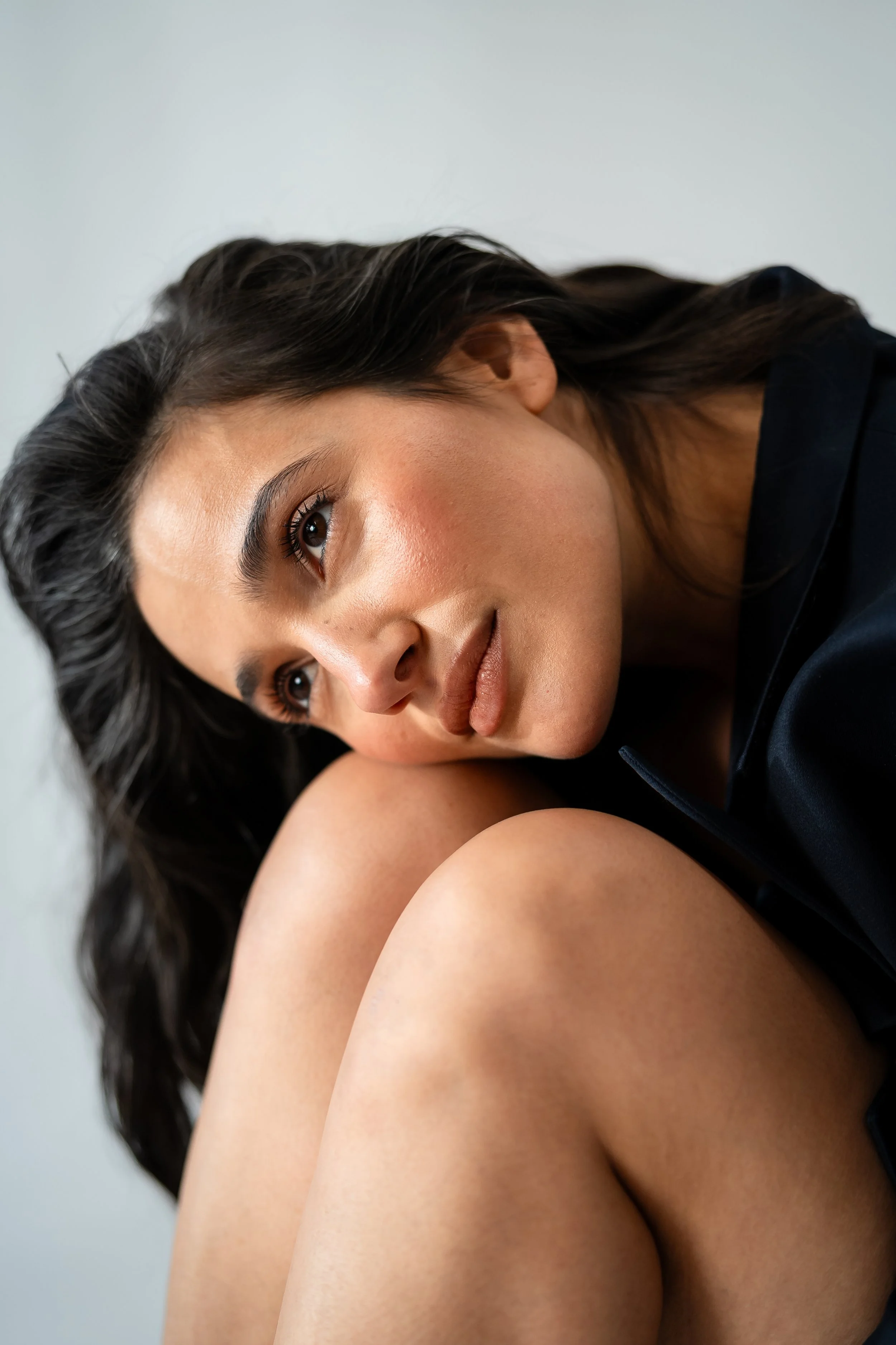 A close-up portrait of a woman with dark hair, resting her head on her knee, looking thoughtfully to the side, with a neutral background.