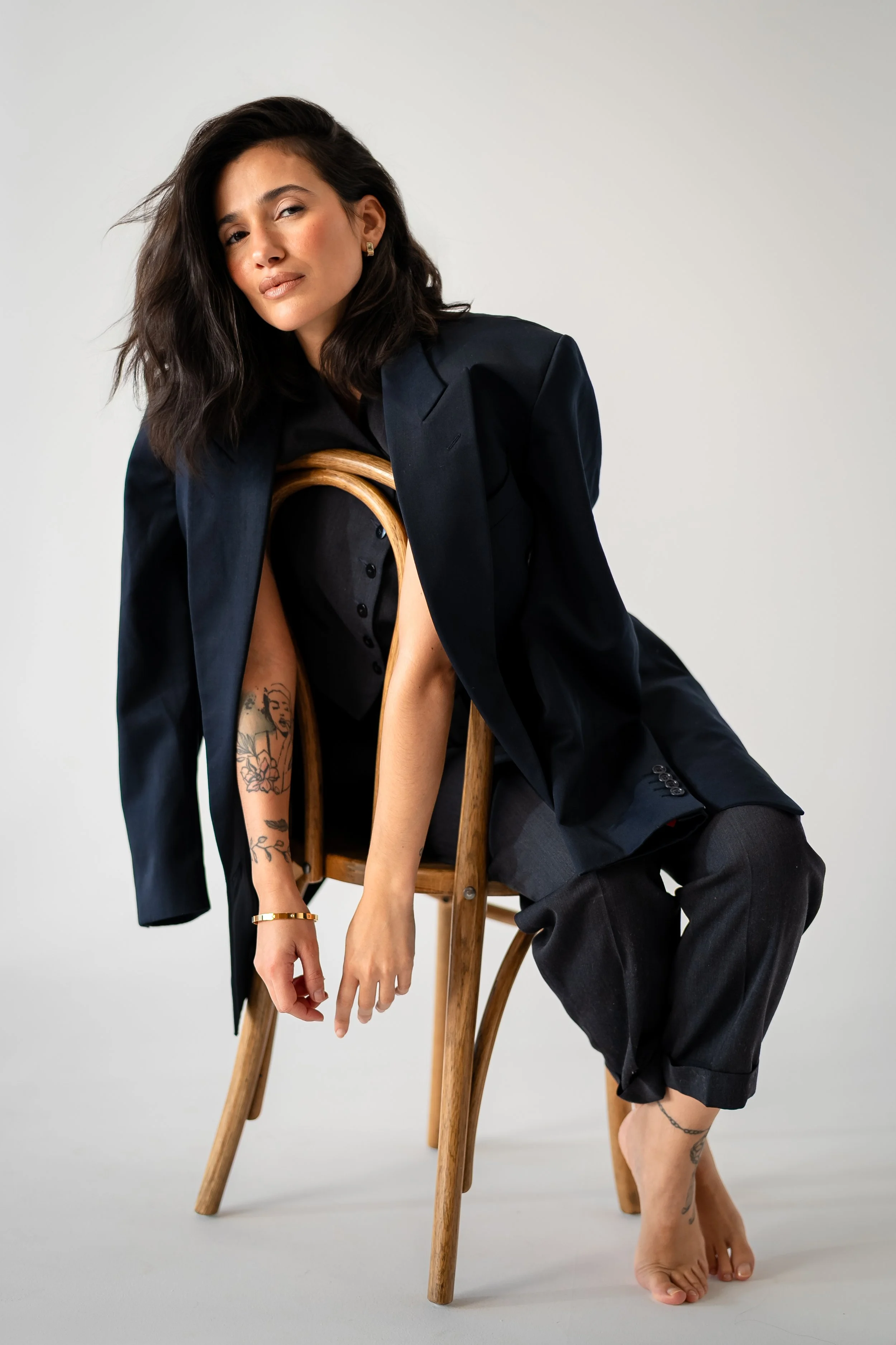 A woman with dark, wavy hair sitting on a wooden chair, wearing a black blazer over a black shirt and gray pants, with tattoos on her arms and ankle, posing against a plain light-colored background.