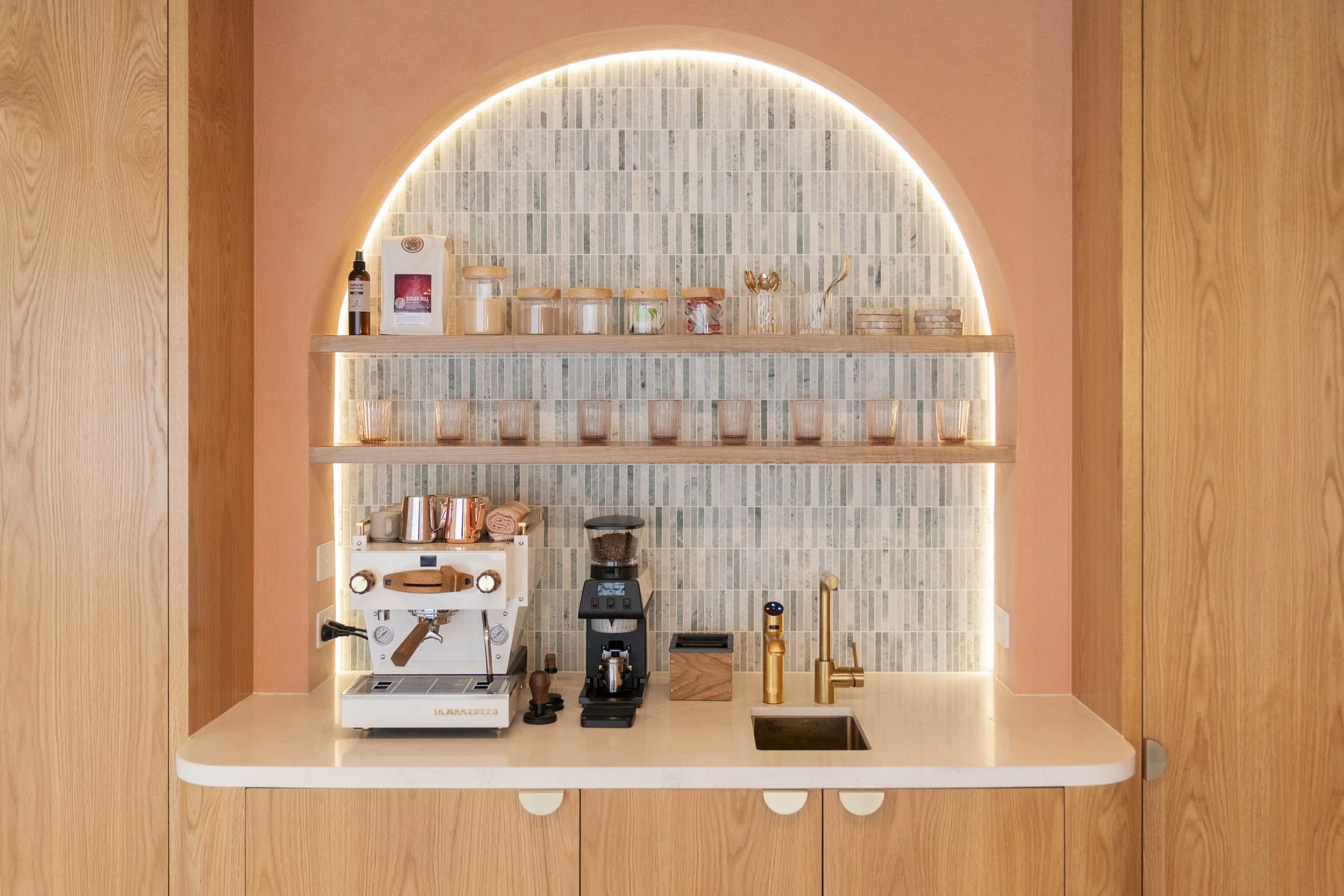 A small coffee station with a white espresso machine, coffee grinder, and gold tap, set against a tiled backsplash with wooden shelves holding jars, glasses, and accessories.