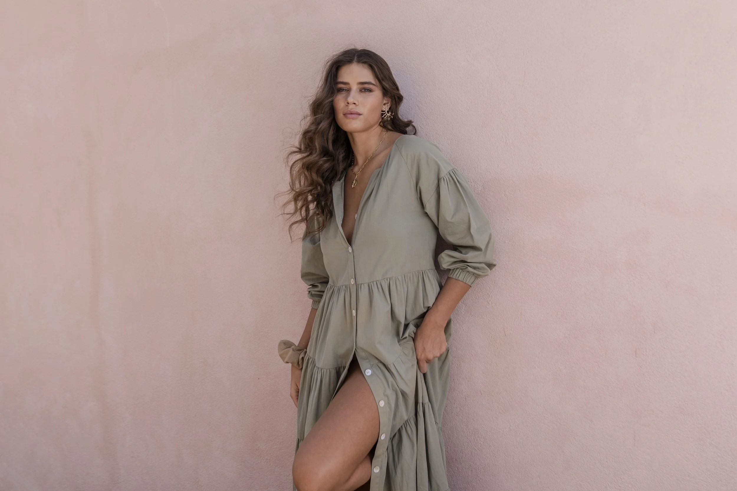 Woman with long, curly brown hair, wearing a beige button-up dress with puffed sleeves and a slit, is standing against a pink wall.