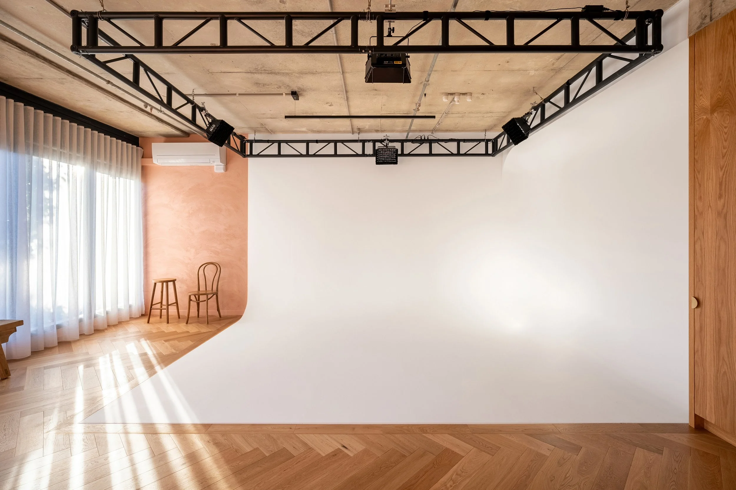 A photography studio with a white curved backdrop, two wooden chairs near a window with sheer curtains, and professional lighting and camera equipment mounted from a ceiling rig.
