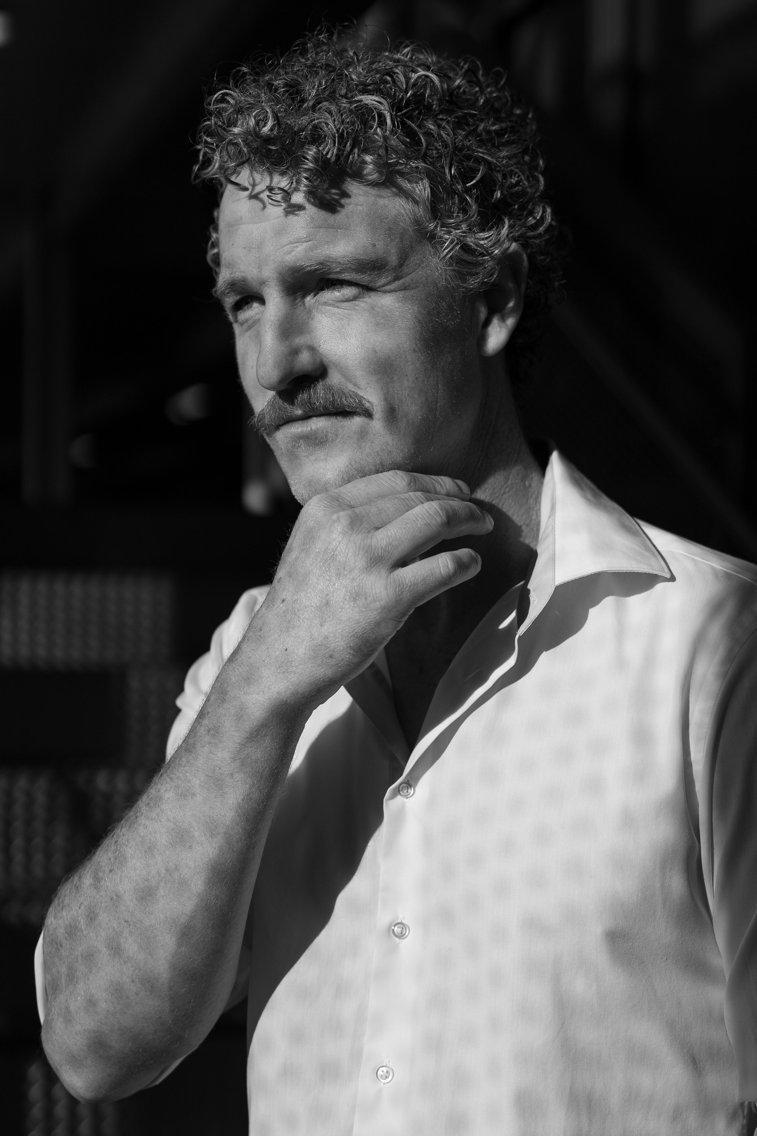 Black and white portrait of a man with curly hair and a mustache, wearing a light-colored shirt, in a contemplative pose outside.