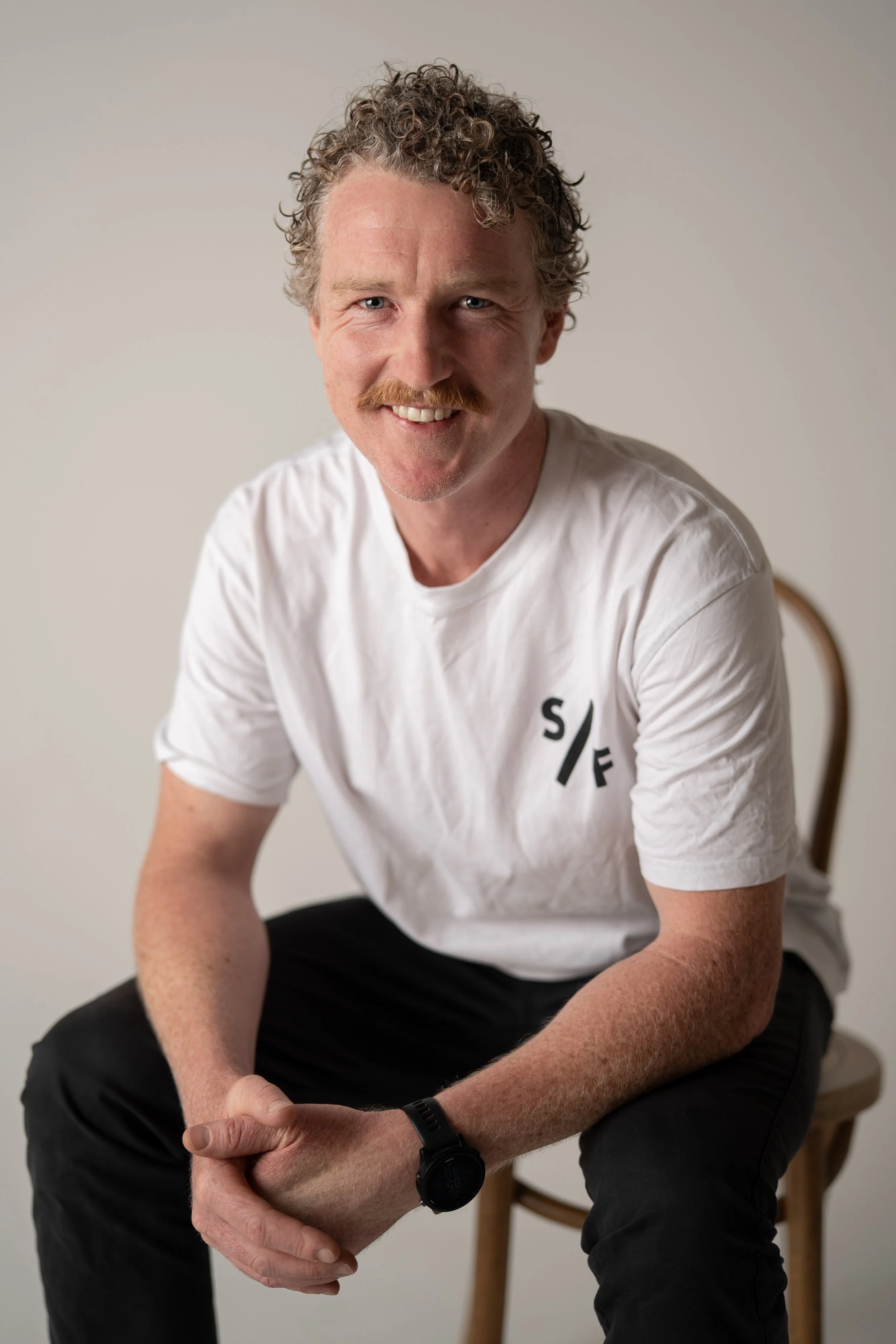 A man with curly hair and a mustache sitting on a wooden chair, smiling, wearing a white t-shirt and black pants, with a black watch on his left wrist, against a plain light background.