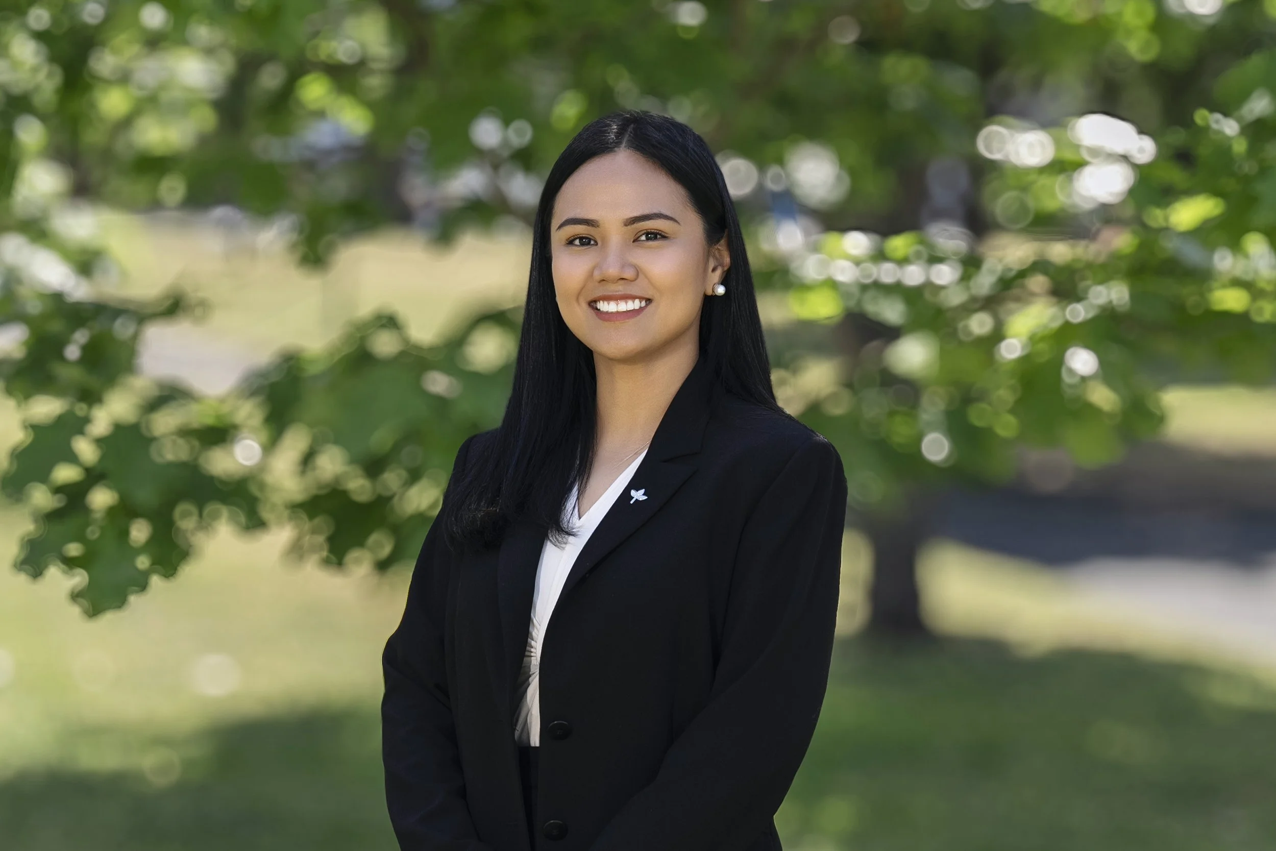A young woman with long black hair, wearing a black blazer, white blouse, and pearl earrings, standing outdoors in front of green trees, smiling at the camera.