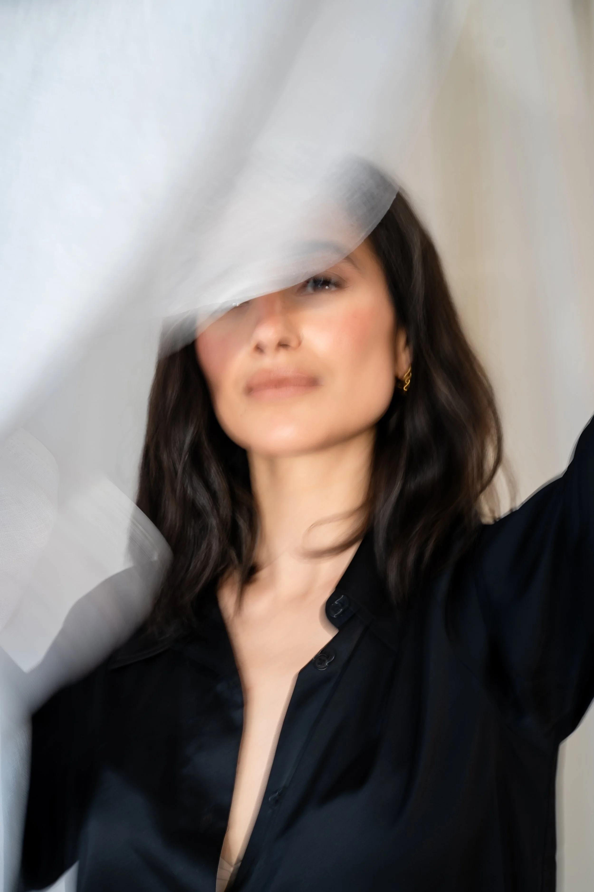 A woman with long dark hair posing with a white fabric or tissue partially covering her face, wearing a black shirt and gold earrings.