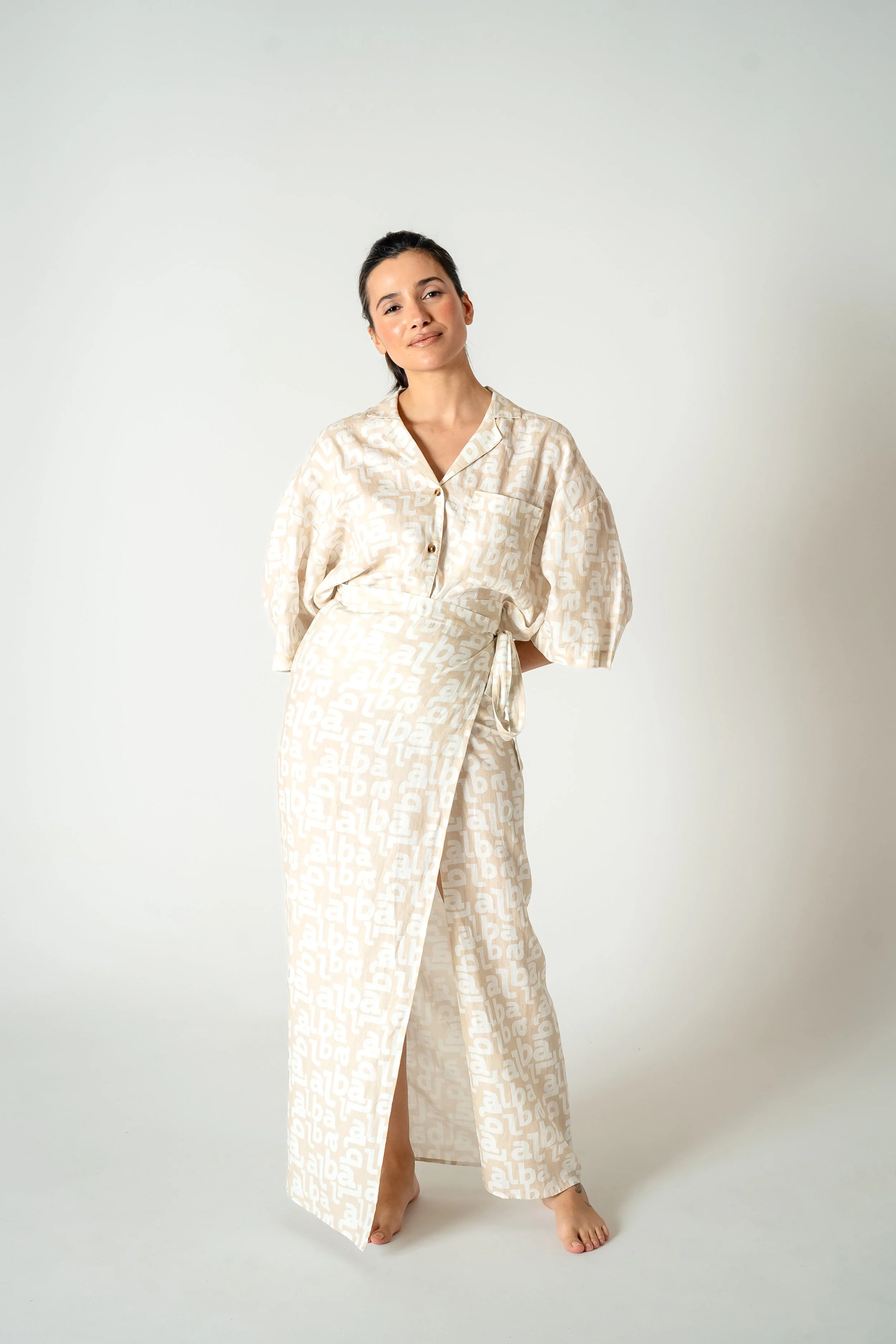 A woman standing barefoot against a plain white background, wearing a matching beige pajama set with a white and beige abstract text pattern, and posing with her head tilted slightly and one hand behind her back.
