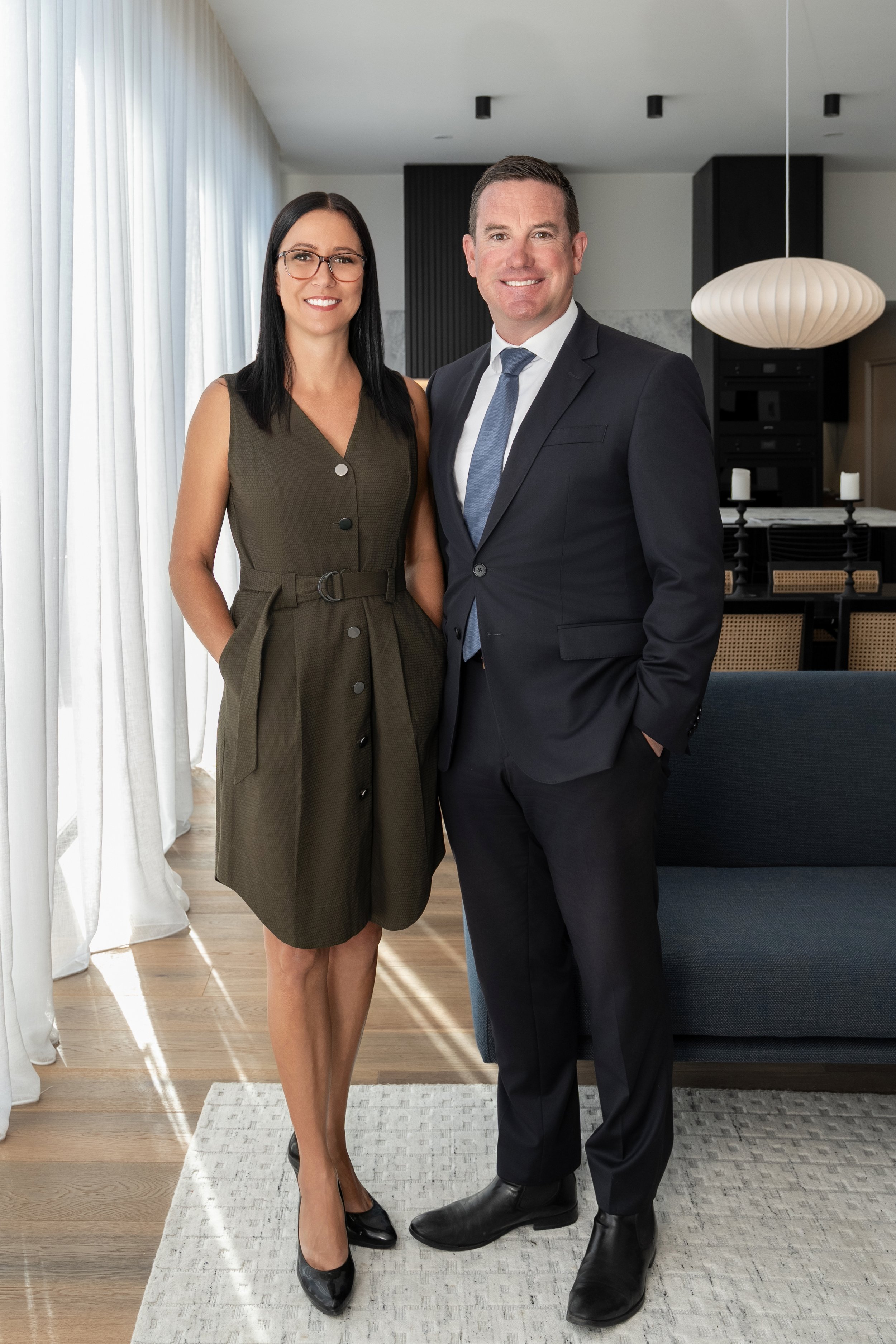 A professionally dressed man and woman standing inside a modern living room, smiling at the camera.